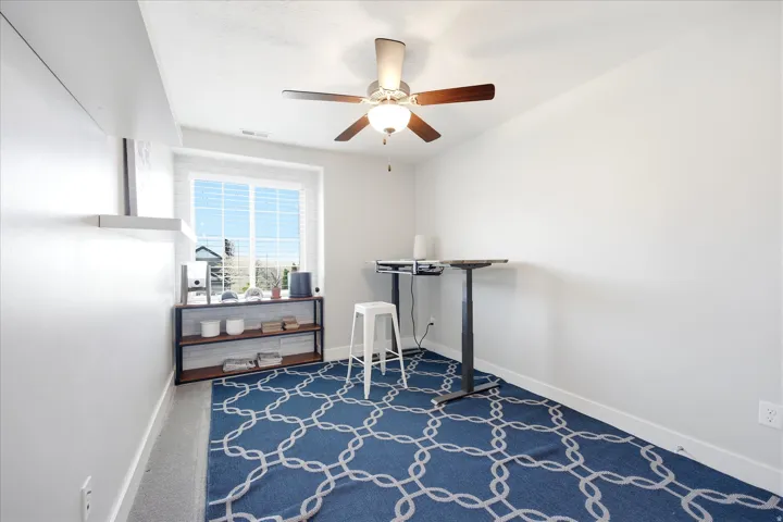 Well-lit room featuring a white-framed window with horizontal blinds, light gray wall-to-wall carpeting, and a ceiling fan with wood-finish blades and integrated lighting