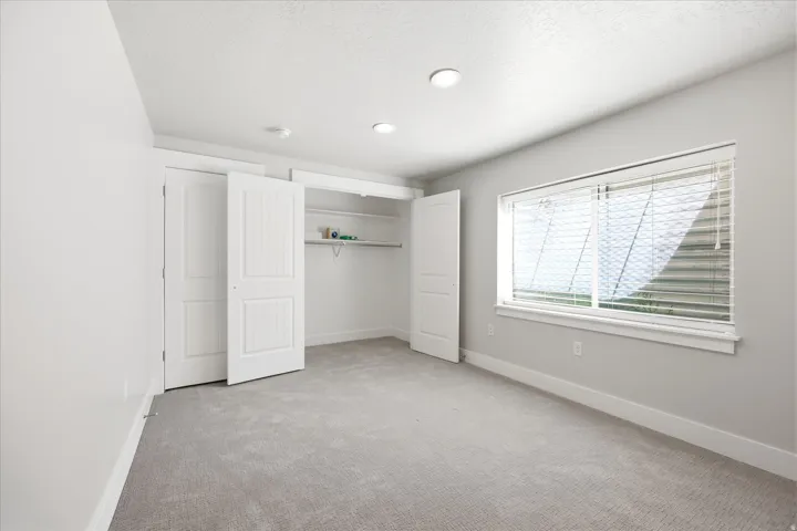 Carpeted room featuring a double-door closet with built-in shelving, a wide window with horizontal blinds, recessed ceiling lighting, and white baseboards