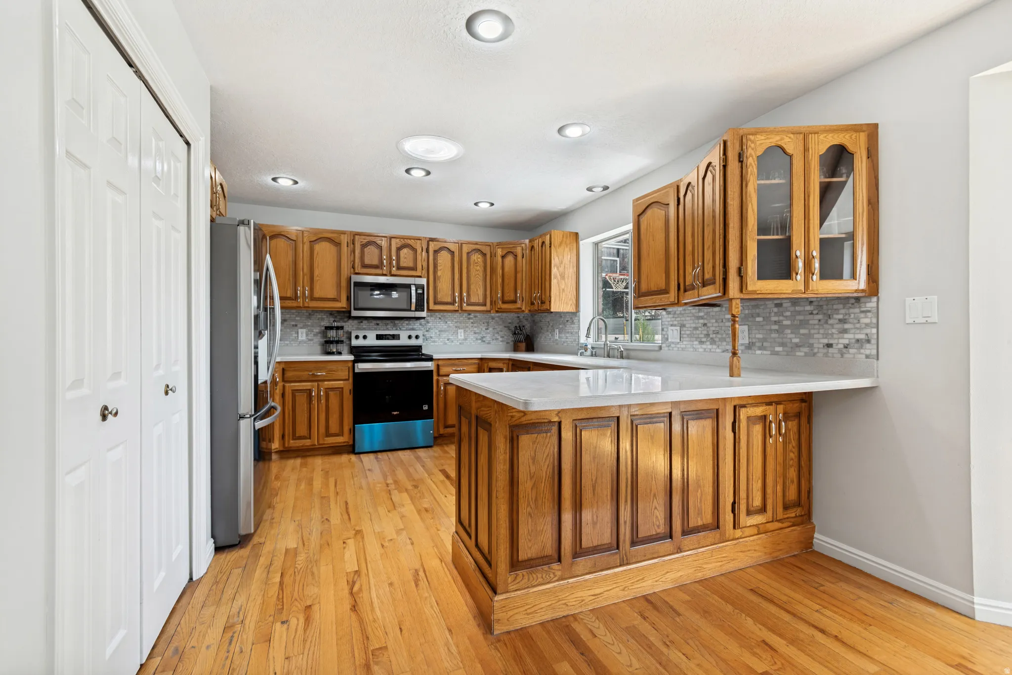 Kitchen featuring honey-toned wood cabinetry, stainless steel appliances, a mosaic tile backsplash, a white countertop, and wood-finish flooring