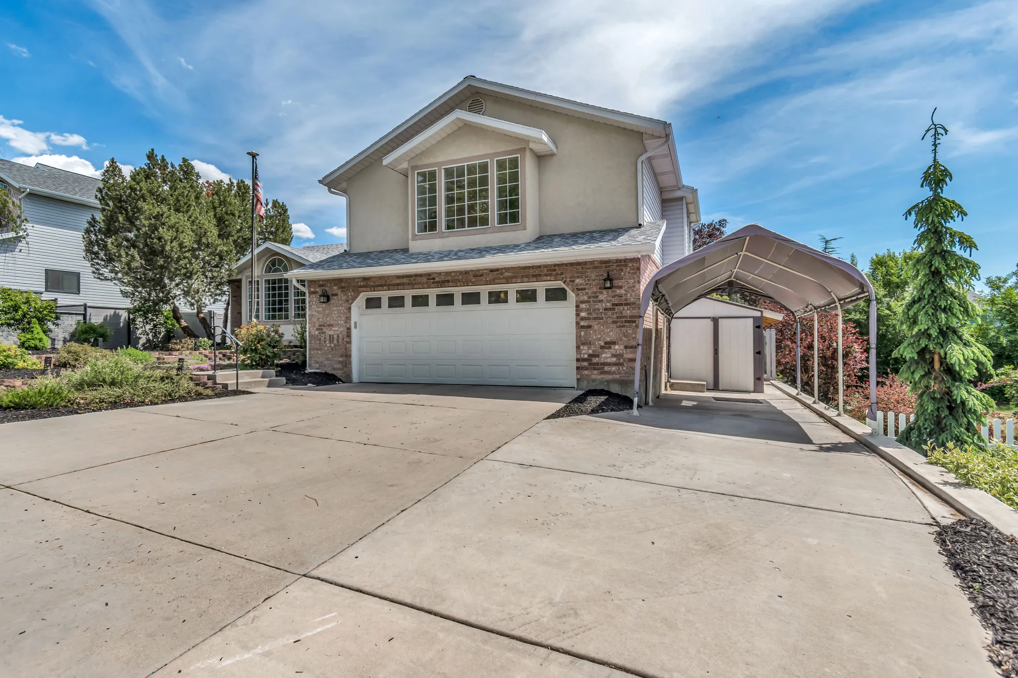 Two-story exterior featuring a brick and stucco facade, a two-car garage, an expansive concrete driveway, a dedicated carport, and tiered landscaping