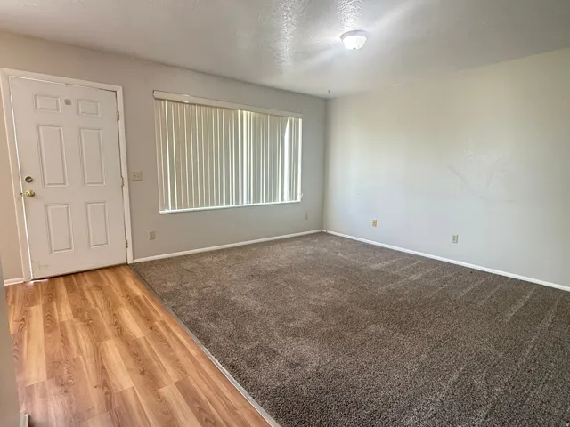 Spacious living area featuring neutral-toned walls, a large window with vertical blinds, and a combination of wood-finish flooring and plush carpeting