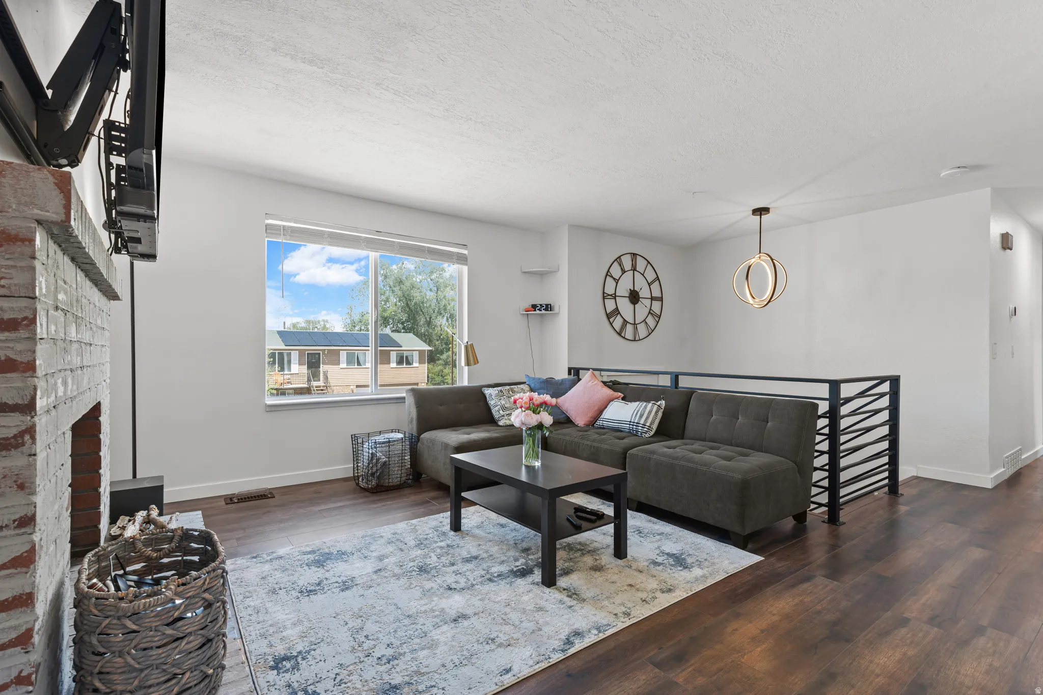 Spacious living area featuring rich wood-finish flooring, a white-painted brick fireplace, a large picture window, a modern pendant light fixture, and a contemporary metal railing