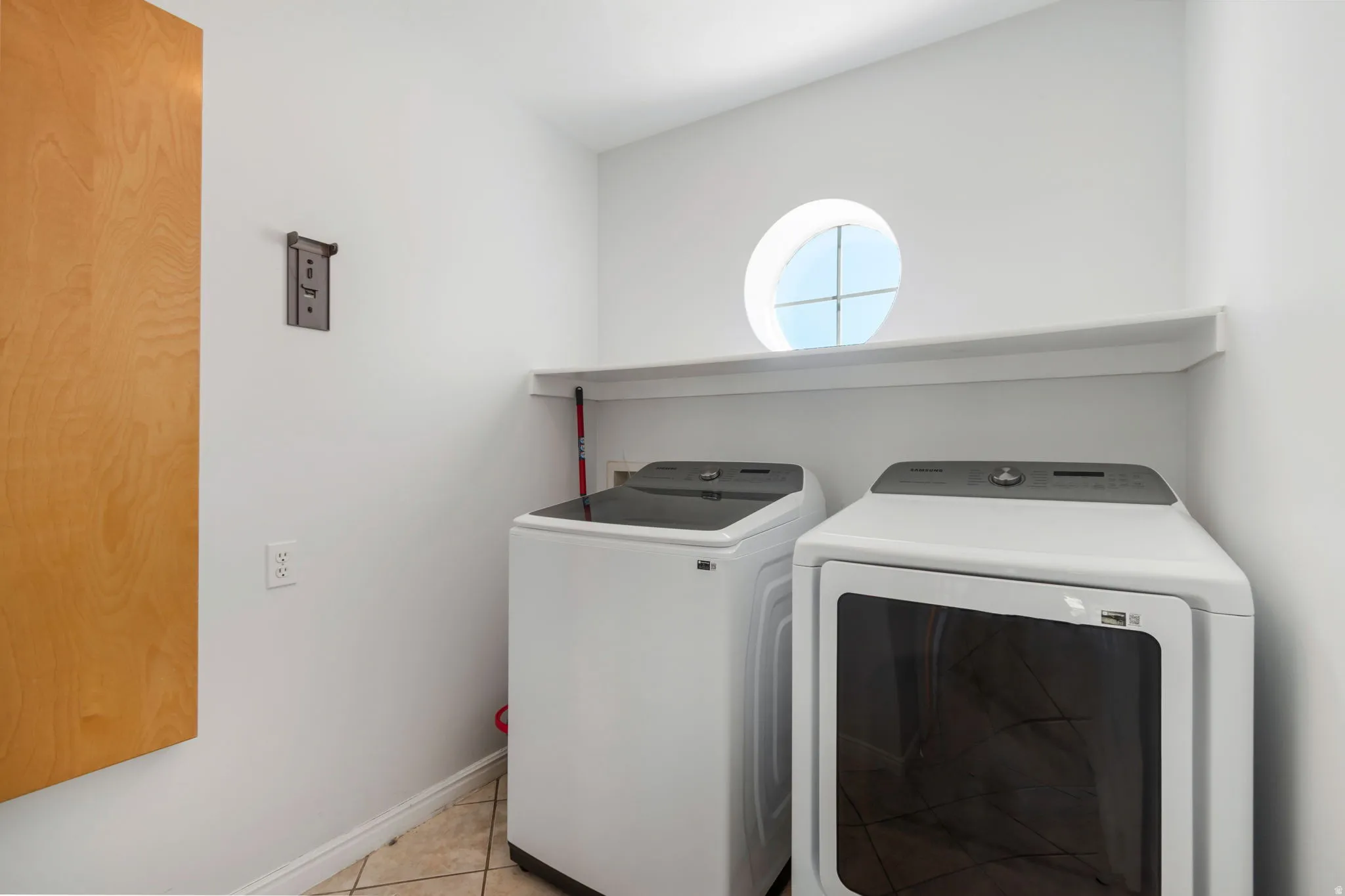 Dedicated laundry space featuring a prominent circular window, tiled flooring, built-in shelving, and light wood-finish cabinetry