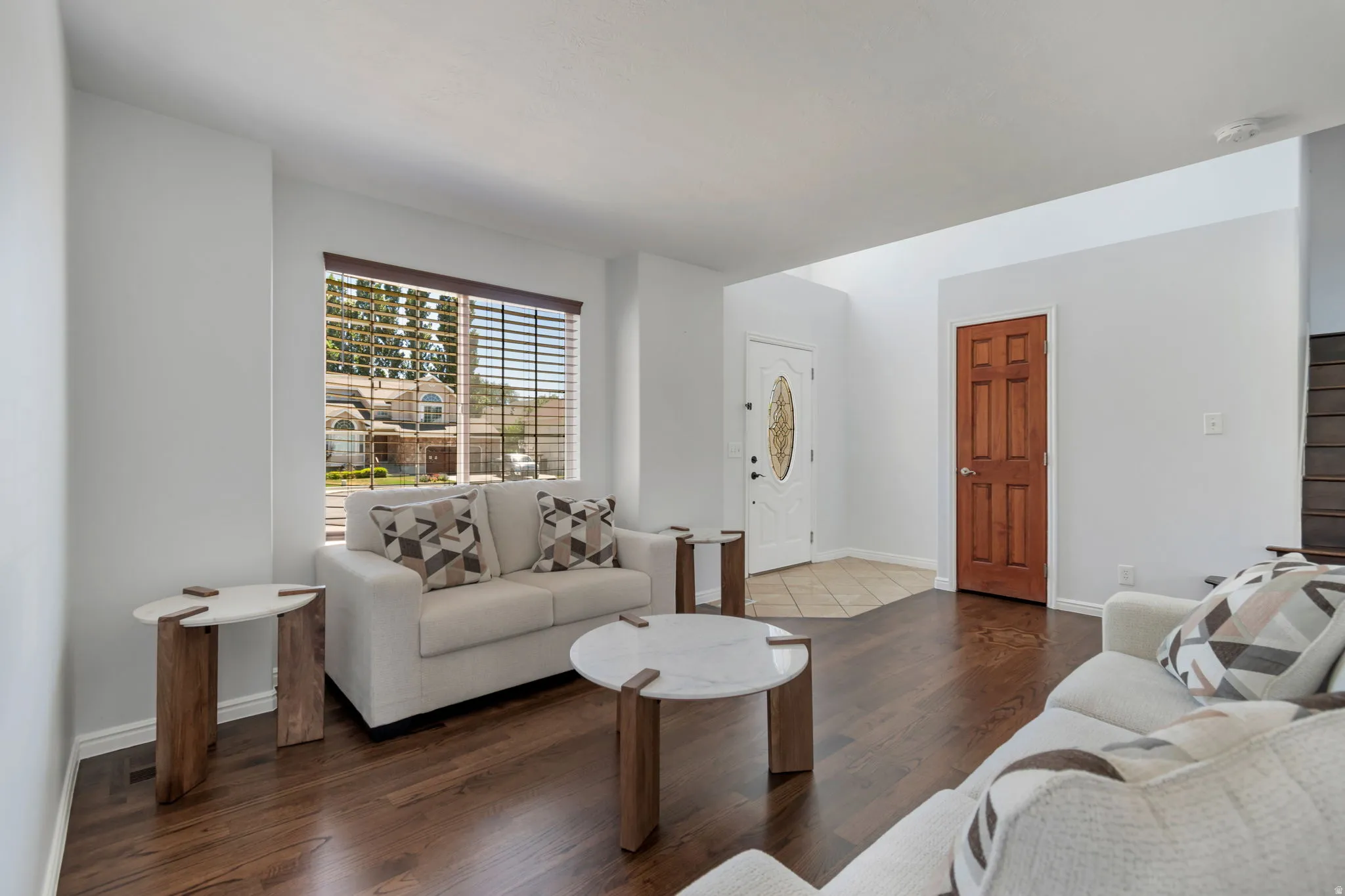 Spacious living area featuring rich wood-finish flooring, a bright window with blinds, and a white front entry door with a decorative glass insert