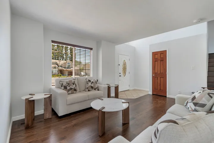 Spacious living area featuring rich wood-finish flooring, a bright window with blinds, and a white front entry door with a decorative glass insert