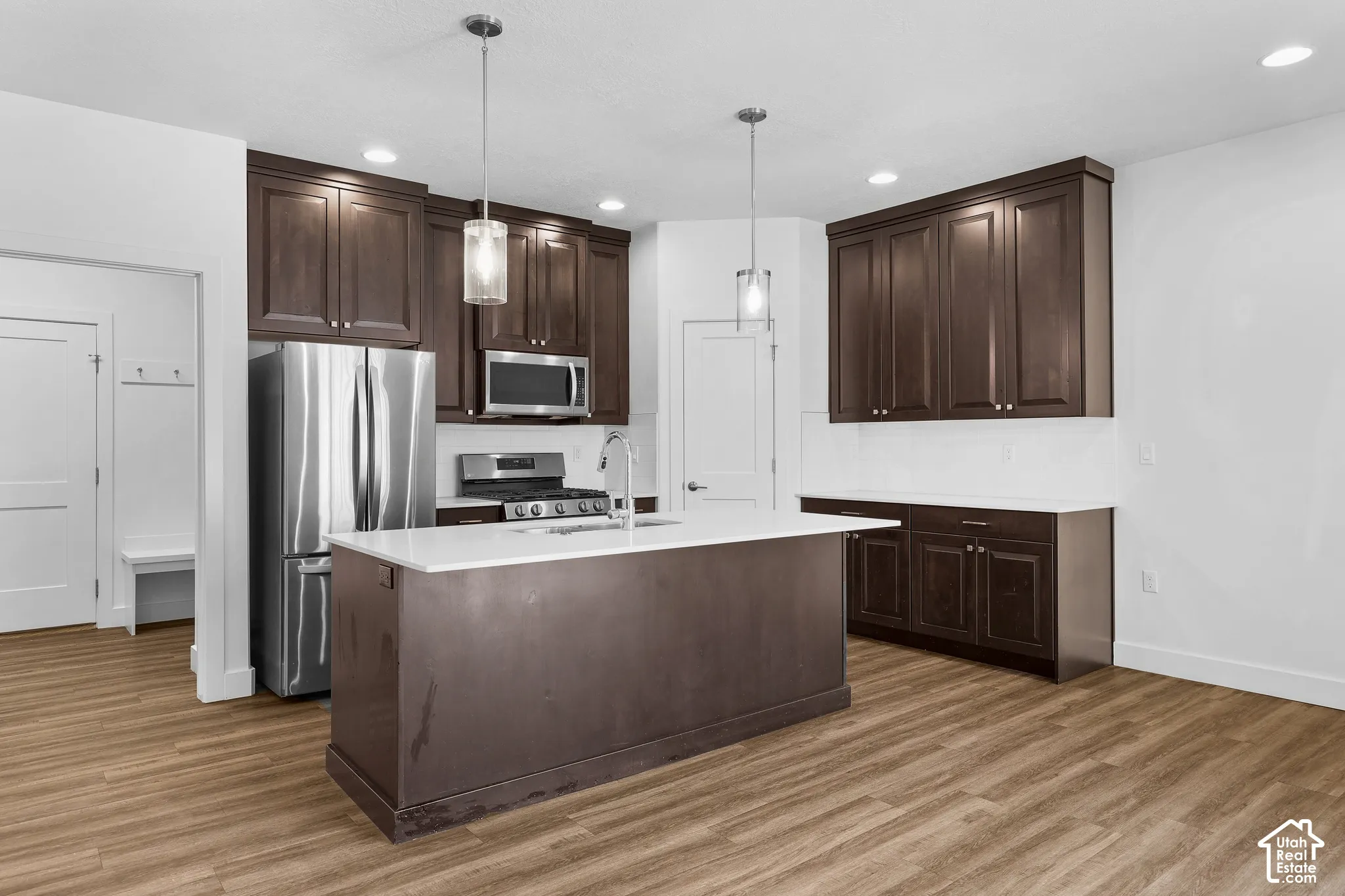 Kitchen featuring dark wood-finish cabinetry, stainless steel appliances, a center island with a sink, pendant lighting, and wood-finish flooring