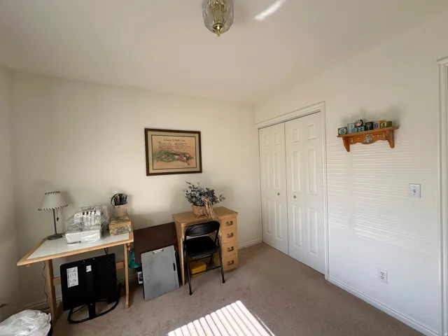 Neutral wall paint and carpeted flooring define this room, featuring a double-door closet with louvered doors, a decorative wall shelf, and a ceiling-mounted light fixture