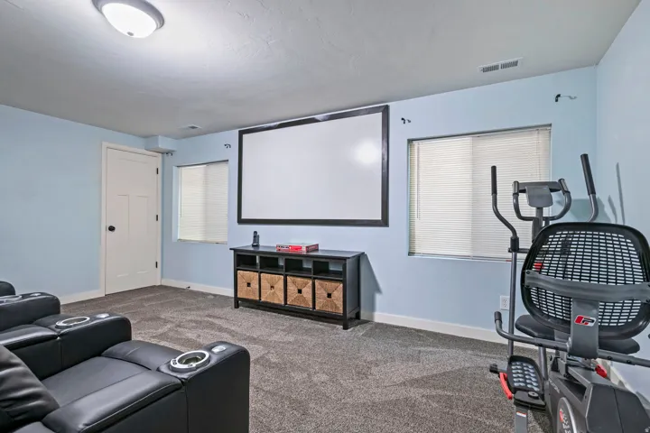 Carpeted recreation room featuring a large projection screen, two windows with blinds, and a built-in media console with storage
