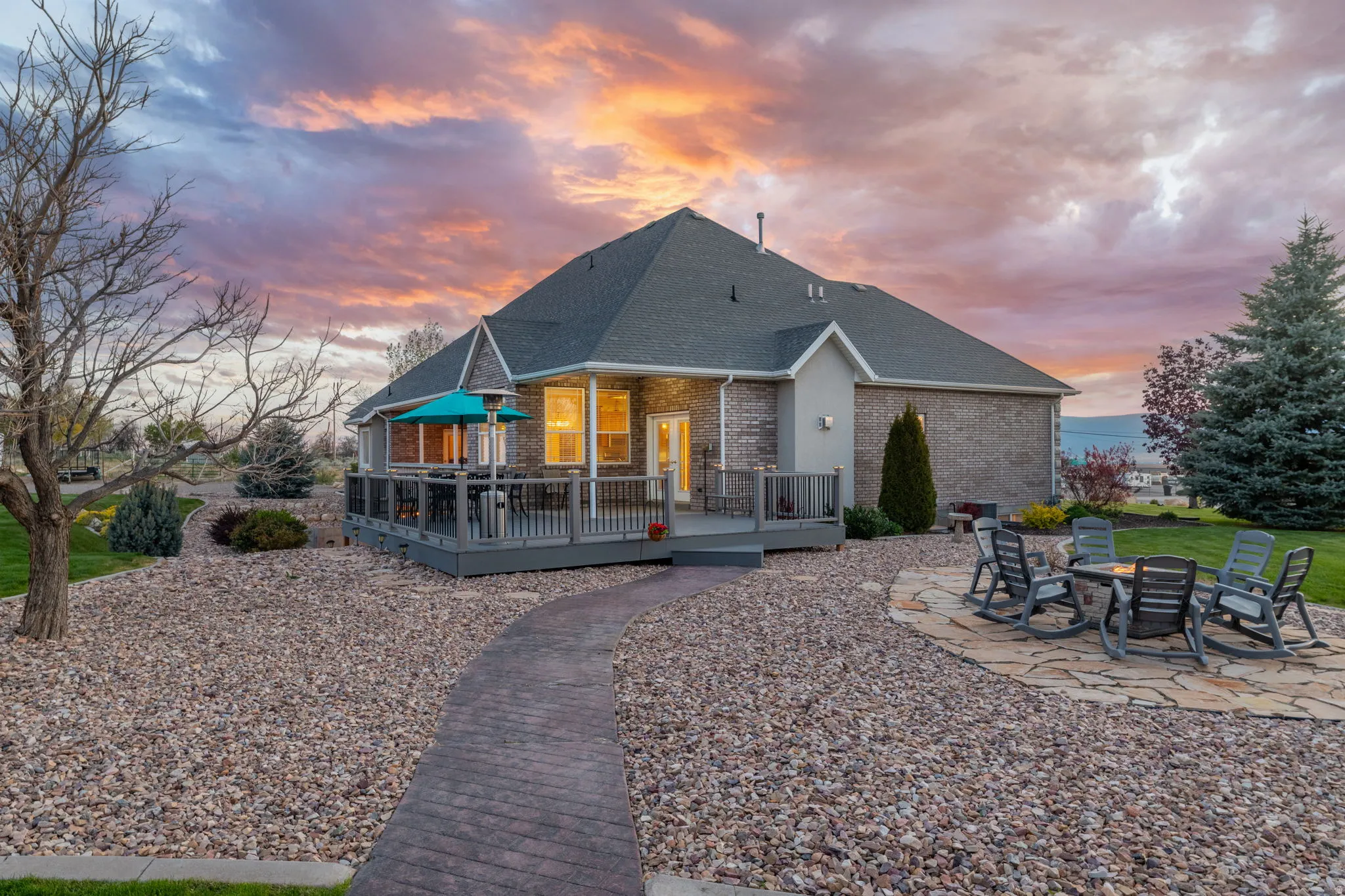 Rear exterior featuring a spacious deck with dark grey railings, a lower-level stone patio, a patterned concrete pathway, and a gravel landscape design