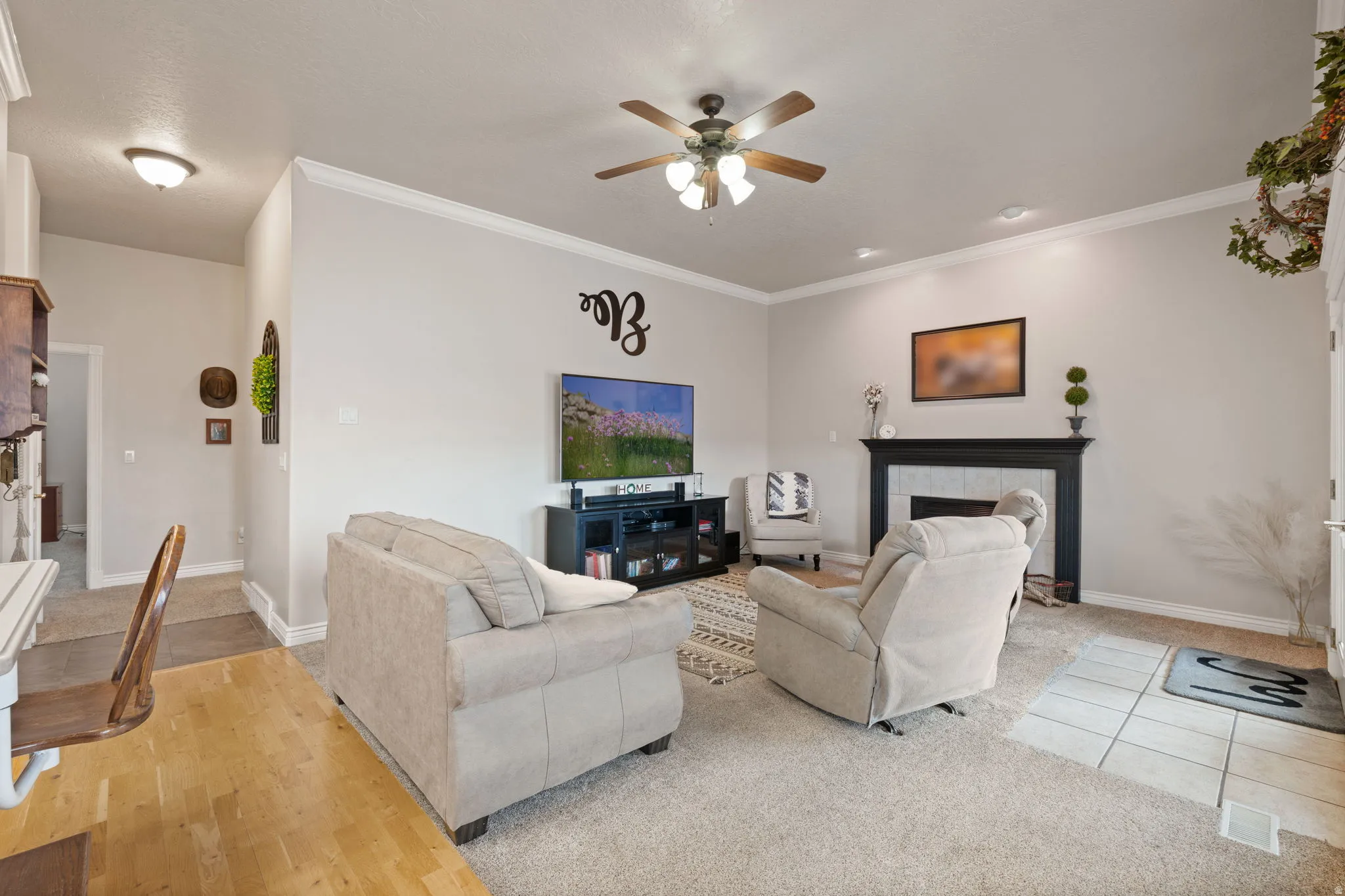 Carpeted living area featuring a central ceiling fan, crown molding, and a fireplace with a dark wood mantel and light-colored tile surround