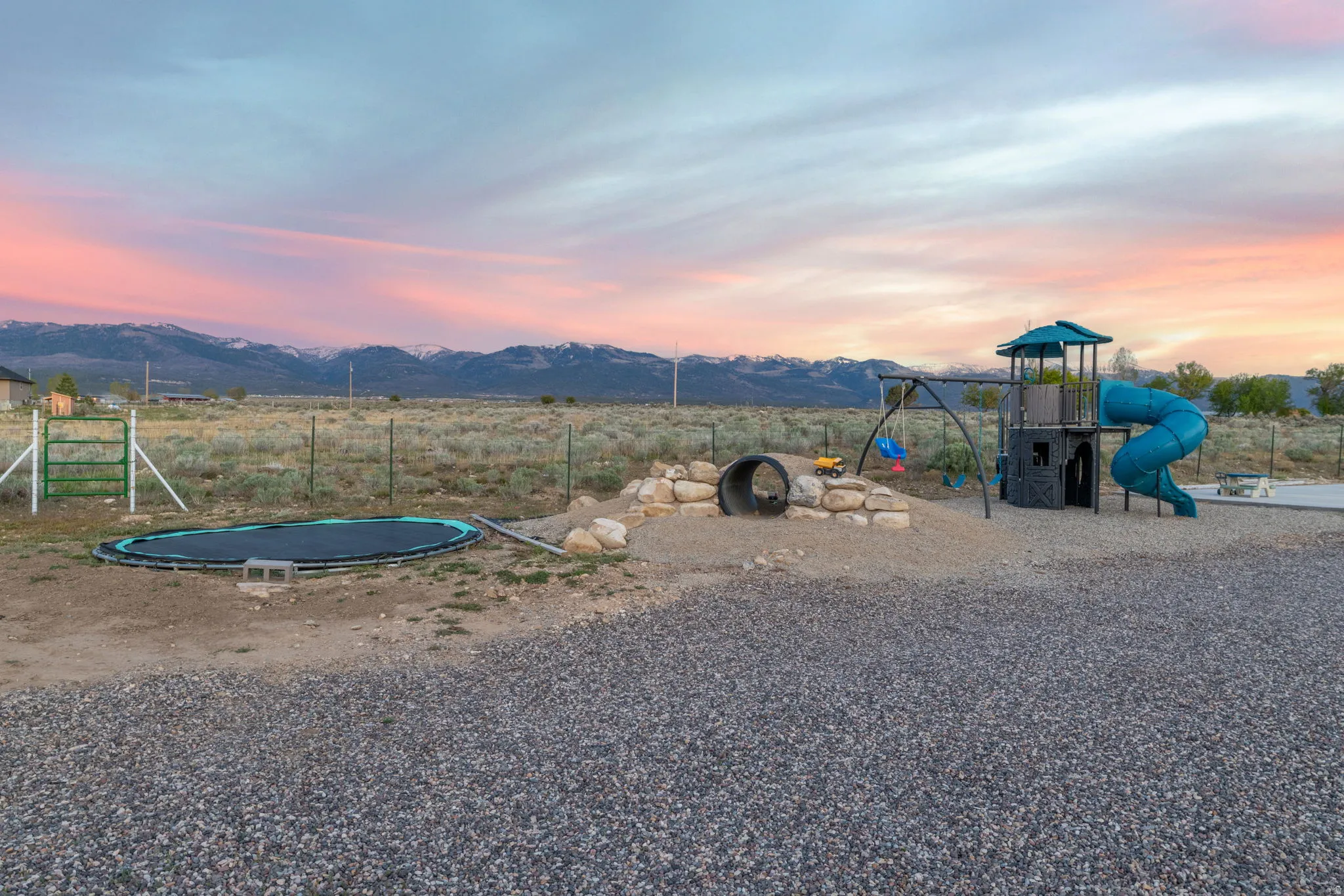 Spacious gravel area featuring a play structure with slide and swings, an in-ground trampoline, and a rock-lined tunnel