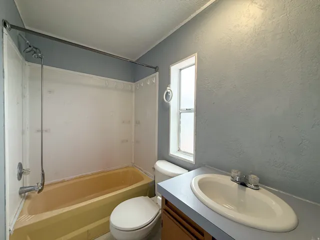 Bathroom featuring a vanity with an integrated sink, a commode, a built-in tub with a shower surround, and a vertical window