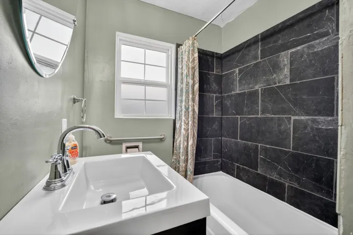 Full bathroom featuring a single white vanity with chrome faucet, a semi-oval wall-mounted mirror, a window with frosted glass, and a tiled shower-tub combination with dark, textured rectangular tiles