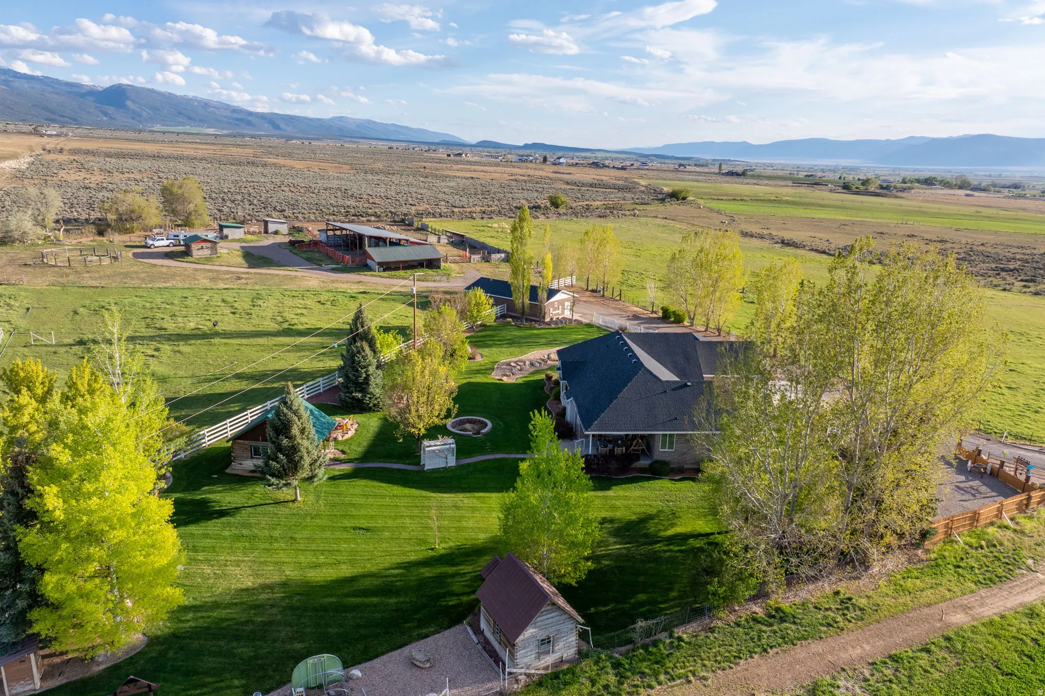Expansive property featuring a residence with a dark shingle roof, a large manicured lawn, several mature trees, a white picket fence, and a distant mountain range