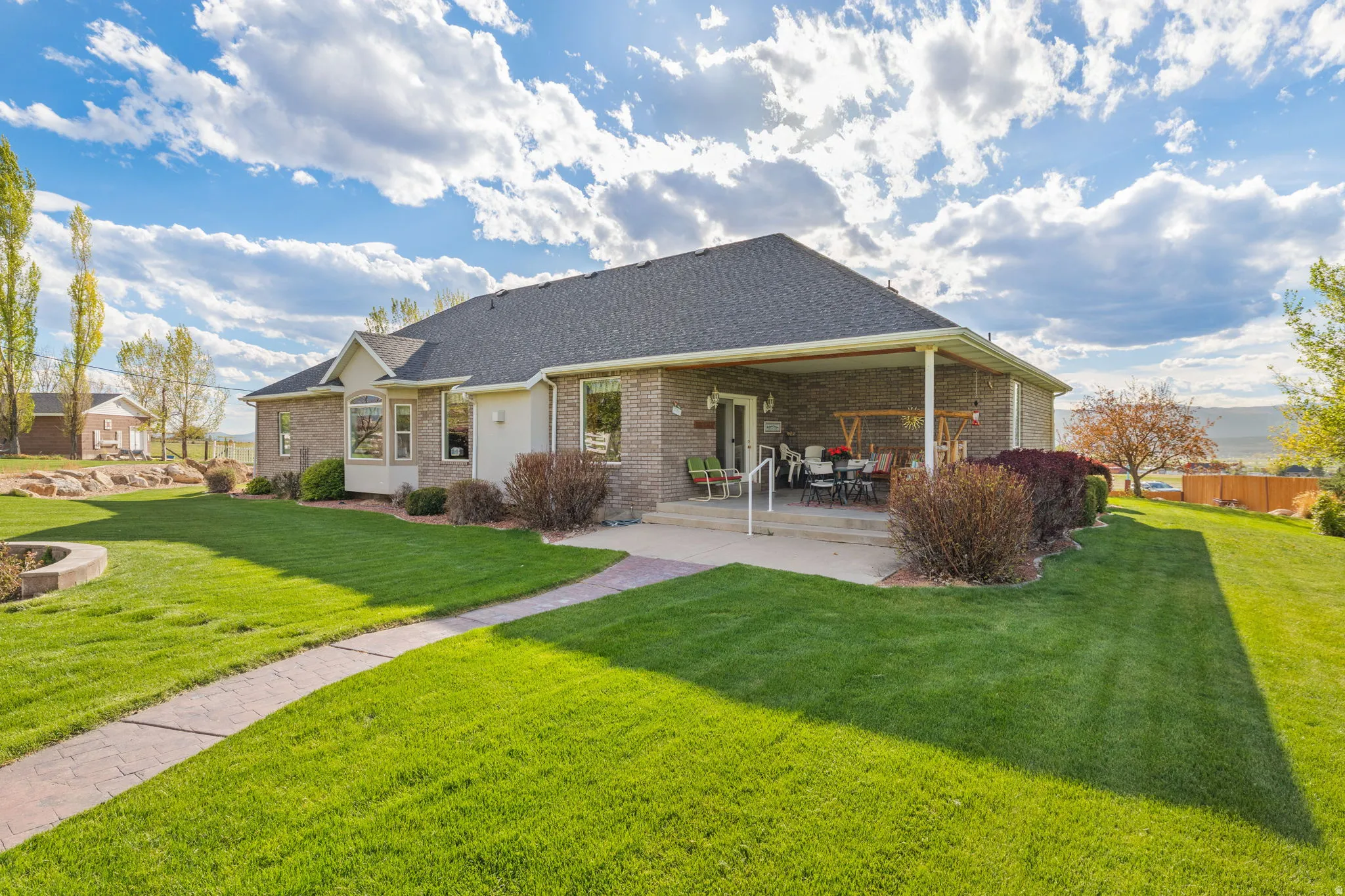 Brick exterior featuring a covered patio, expansive lawn, mature shrubs, and a paved pathway