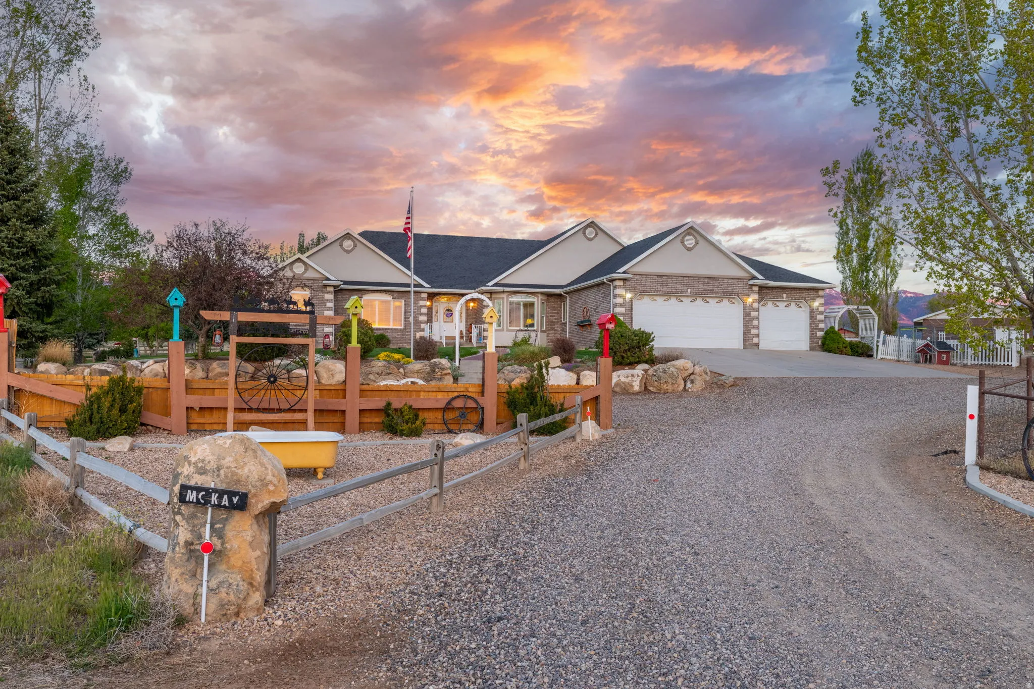 Brick exterior featuring a two-car garage, gravel driveway, and a wooden fence with stone accents