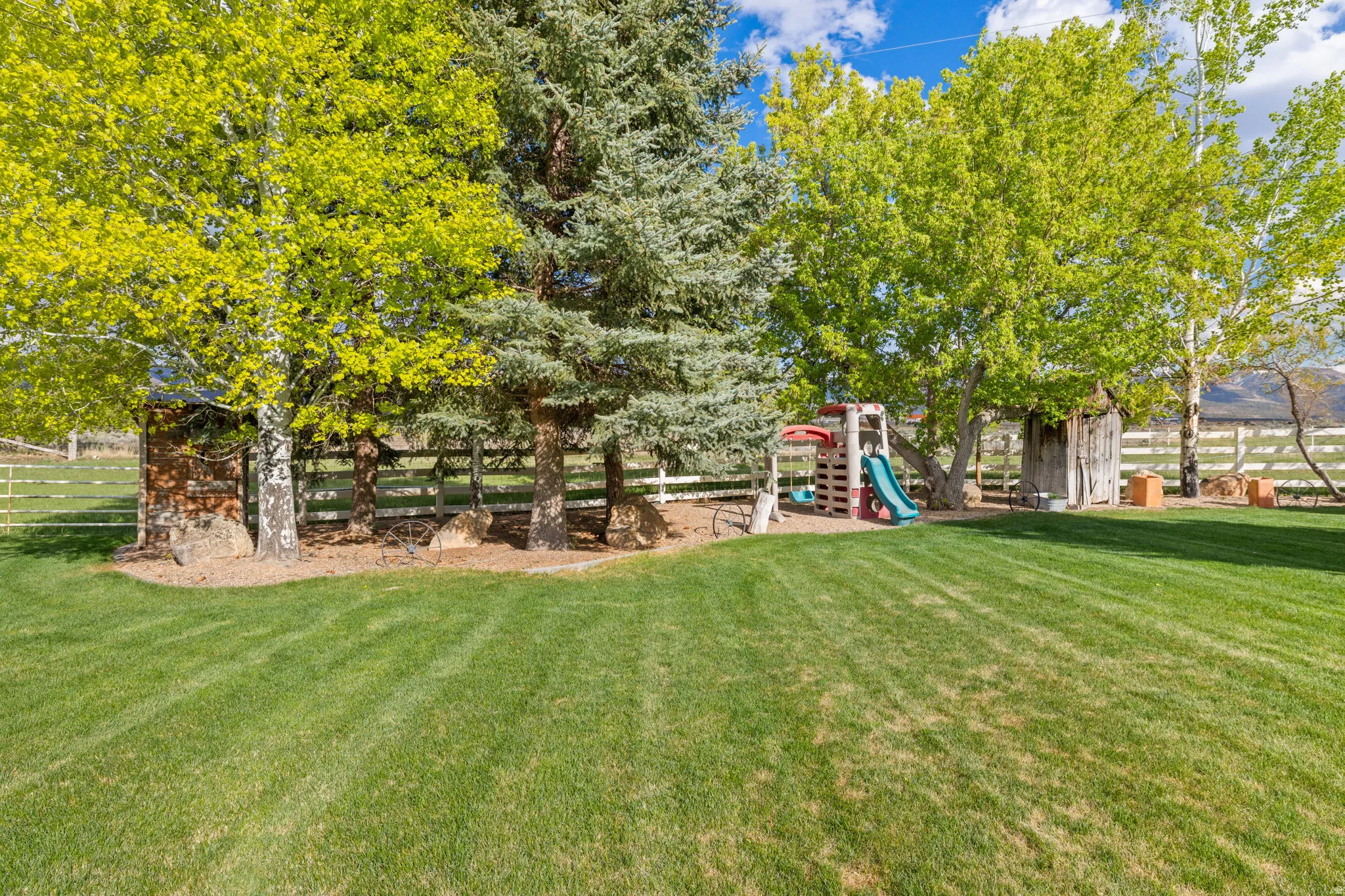 Expansive green lawn surrounded by mature trees, featuring a white perimeter fence and a small wooden shed