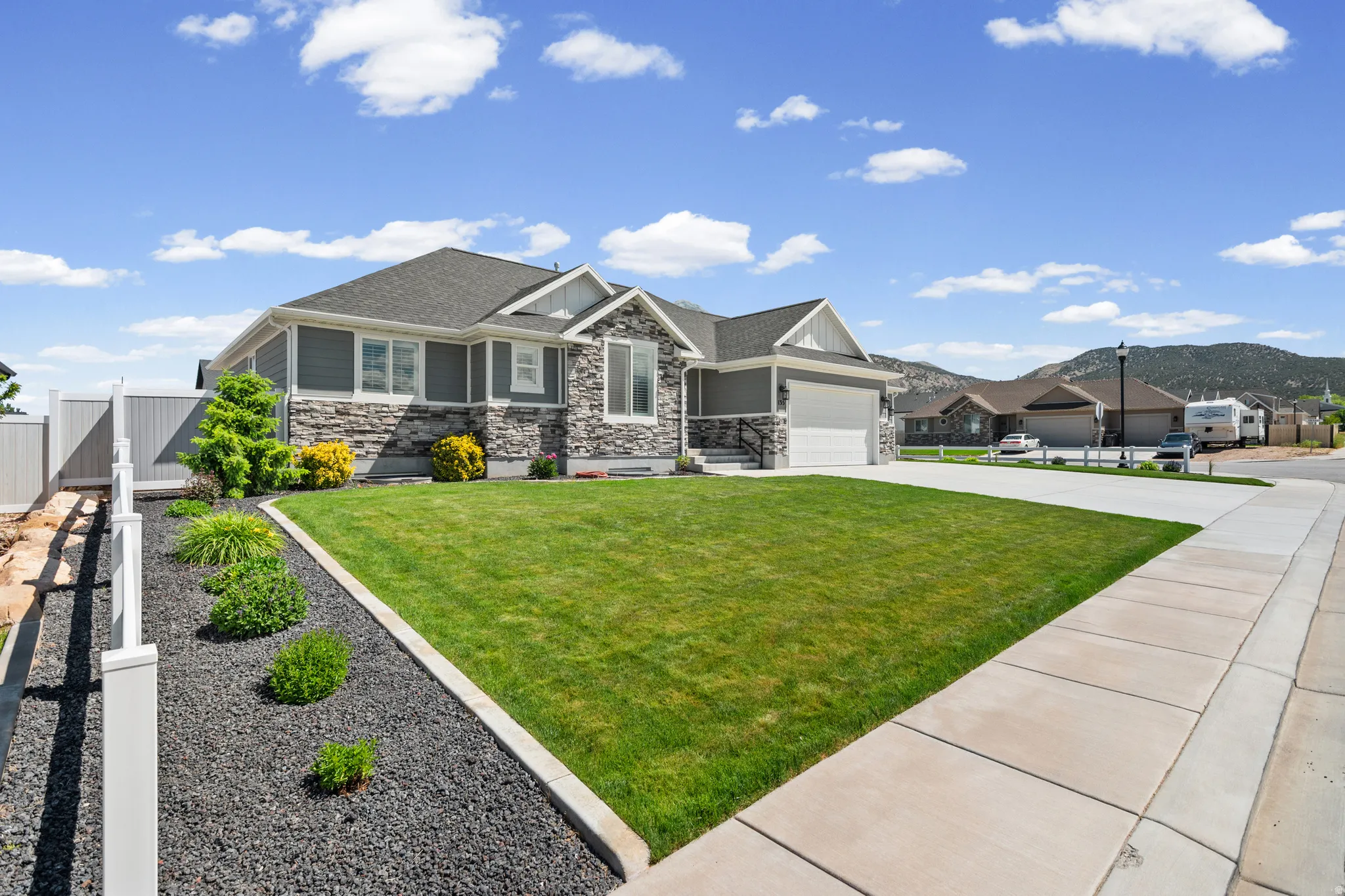 Contemporary residence featuring a stone and siding exterior, a two-car garage, a well-maintained front lawn, and xeriscape landscaping with a white privacy fence