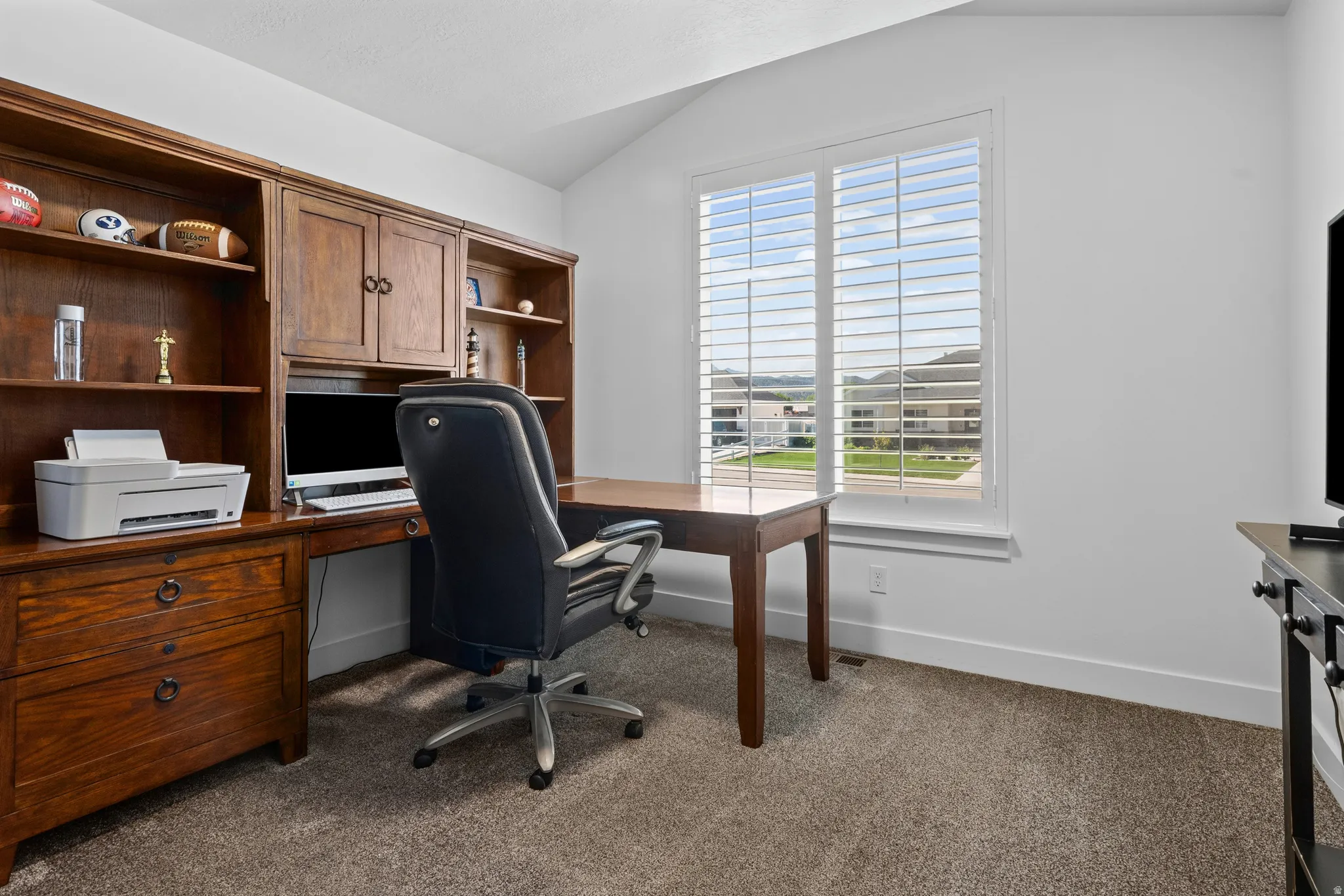 Built-in wood cabinetry with integrated desk space, a large window with plantation shutters, vaulted ceiling, and neutral-tone carpet flooring