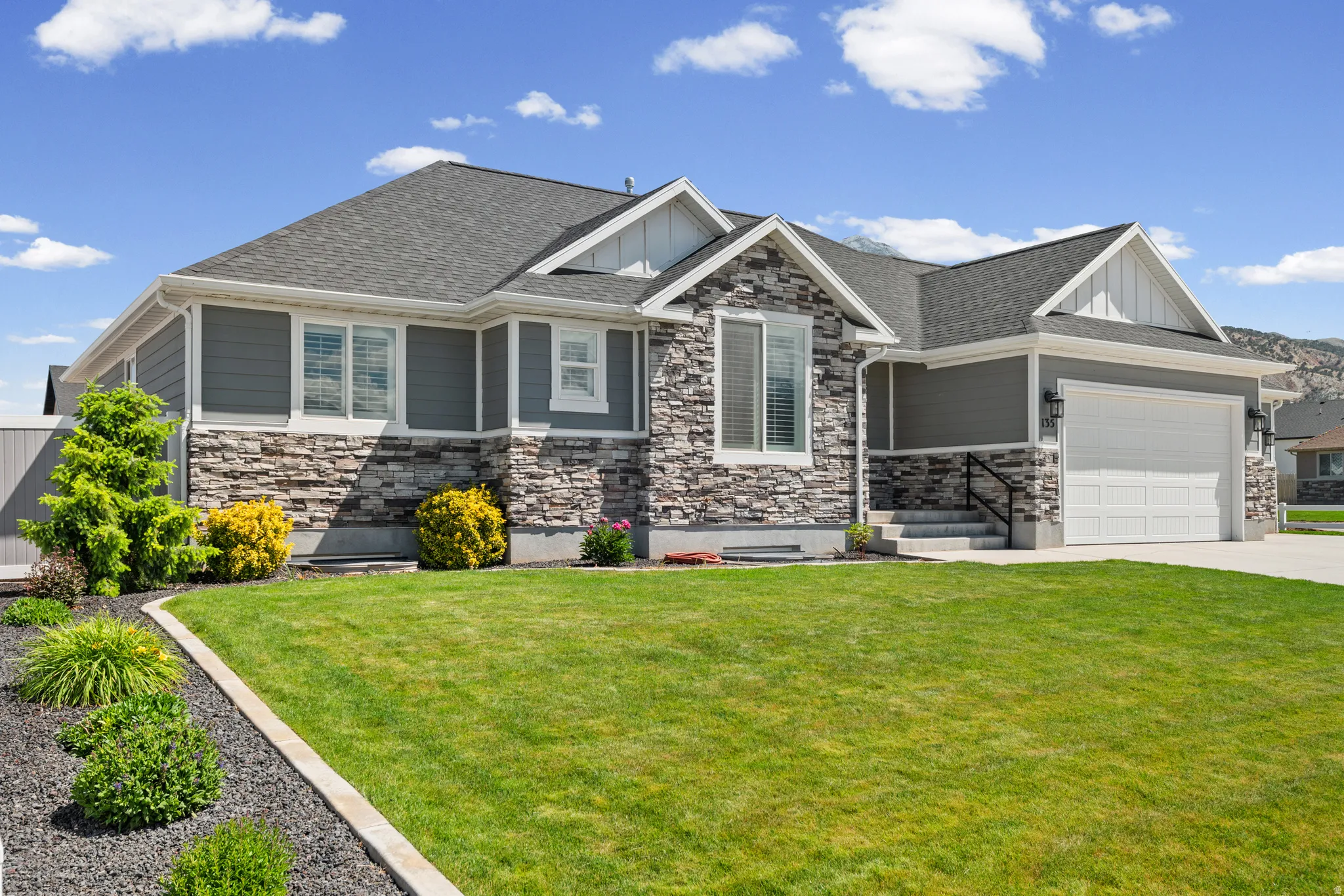 Contemporary facade featuring gray horizontal siding, stacked stone accents, a dark shingle roof, a two-car garage, and established landscaping with a concrete curb and dark rock bedding