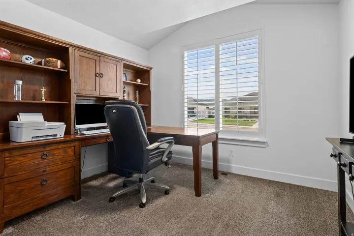 Built-in wood cabinetry with integrated desk space, a large window with plantation shutters, vaulted ceiling, and neutral-tone carpet flooring