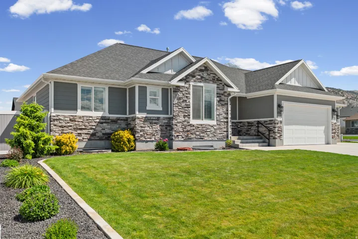 Contemporary facade featuring gray horizontal siding, stacked stone accents, a dark shingle roof, a two-car garage, and established landscaping with a concrete curb and dark rock bedding