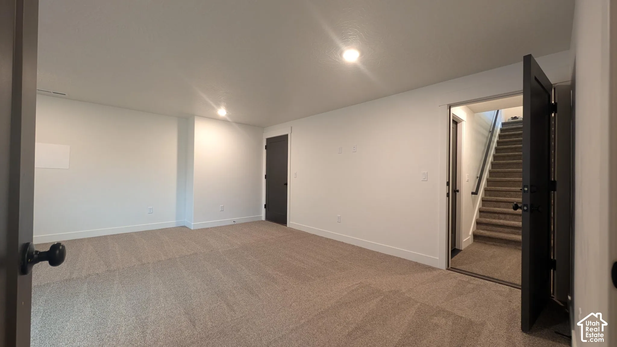 Spacious carpeted room with white walls, recessed lighting, and a dark-paneled door leading to a carpeted staircase with a black handrail