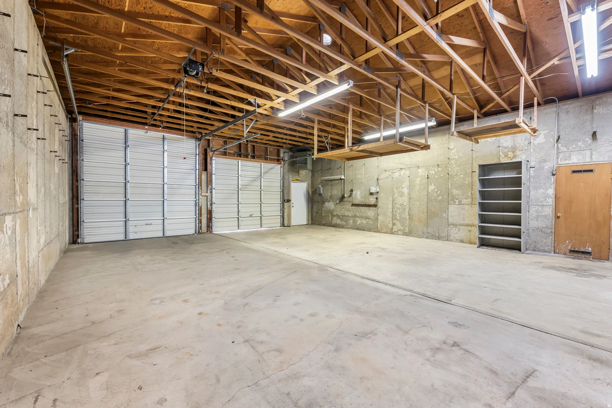 Spacious interior featuring two large roll-up doors, exposed wood beam ceiling, concrete flooring, built-in shelving, and overhead fluorescent lighting