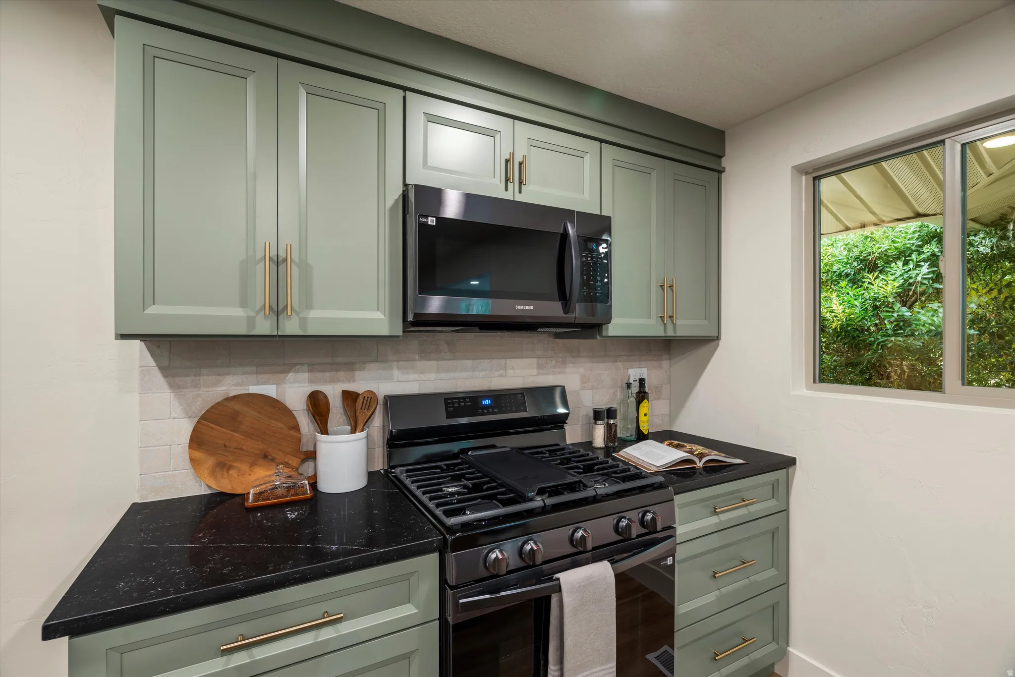 Kitchen featuring sage green cabinetry with gold-tone hardware, black countertop surfaces, stainless steel range, and an over-the-range microwave