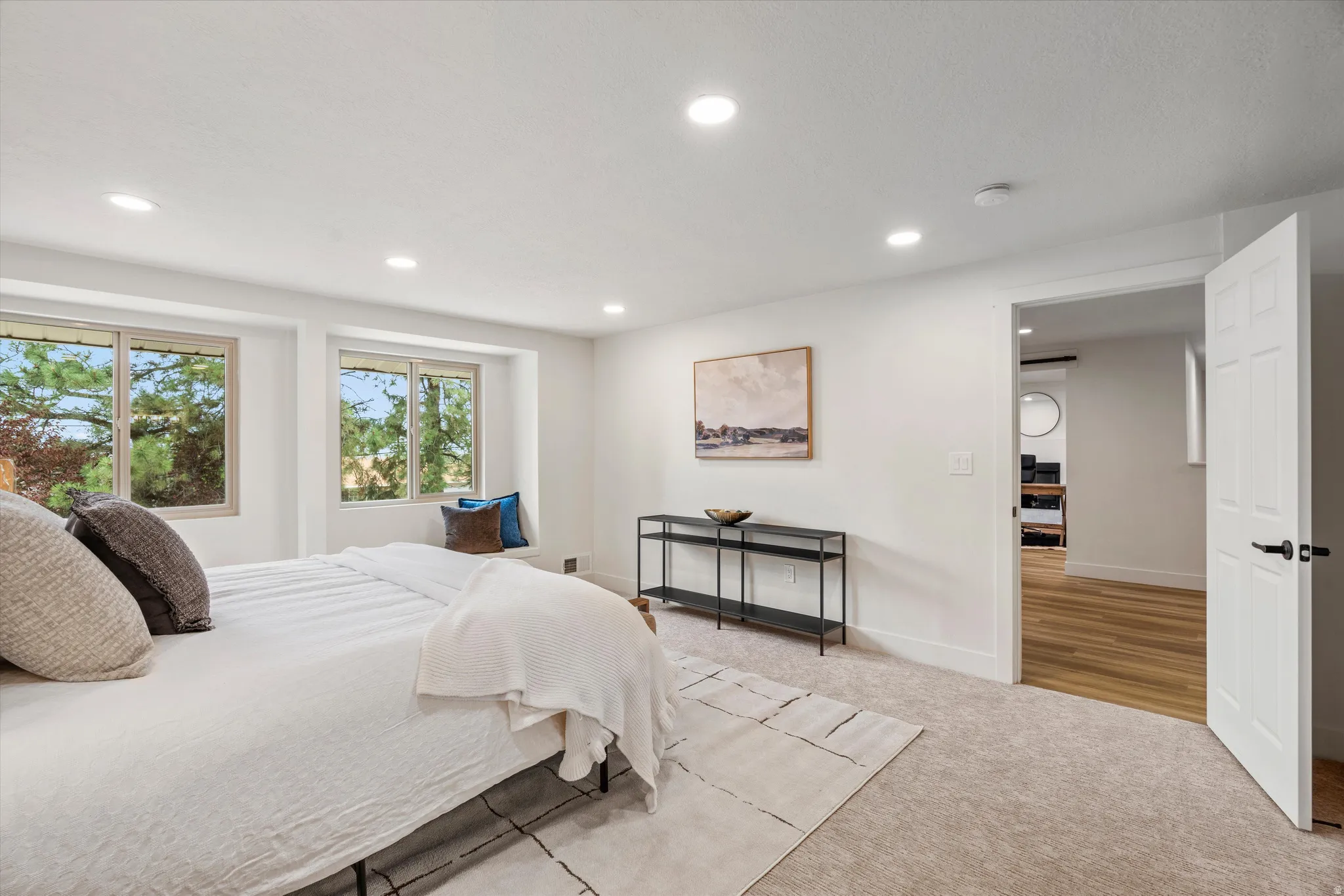 Recessed lighting illuminates this bright bedroom featuring a window seat, light-toned carpeting, and white trim