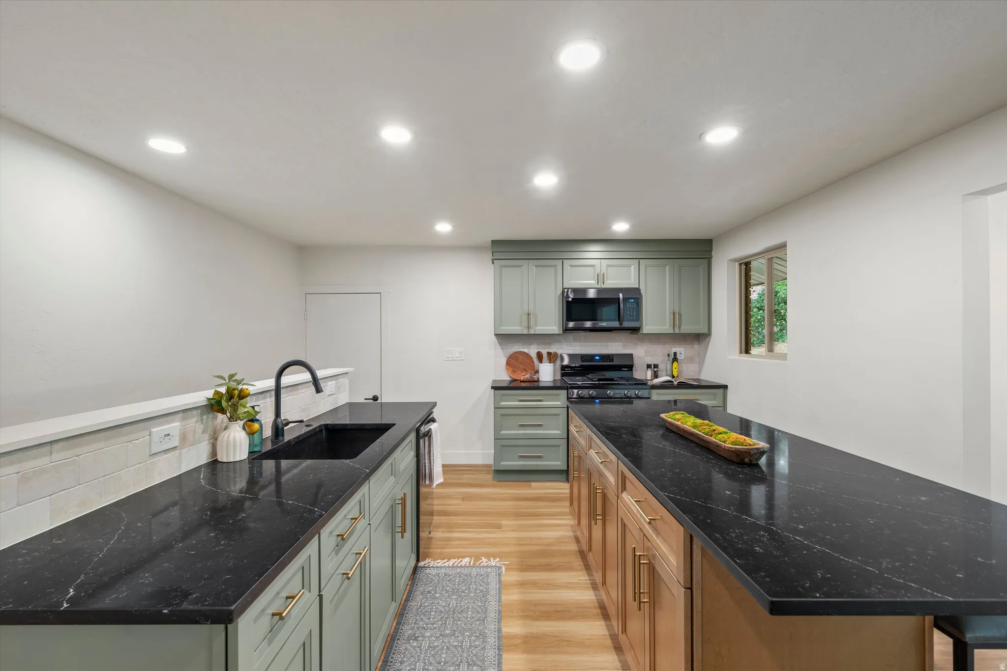 Modern kitchen featuring ample dark countertops, sage green cabinetry with brass hardware, and wood-finish flooring