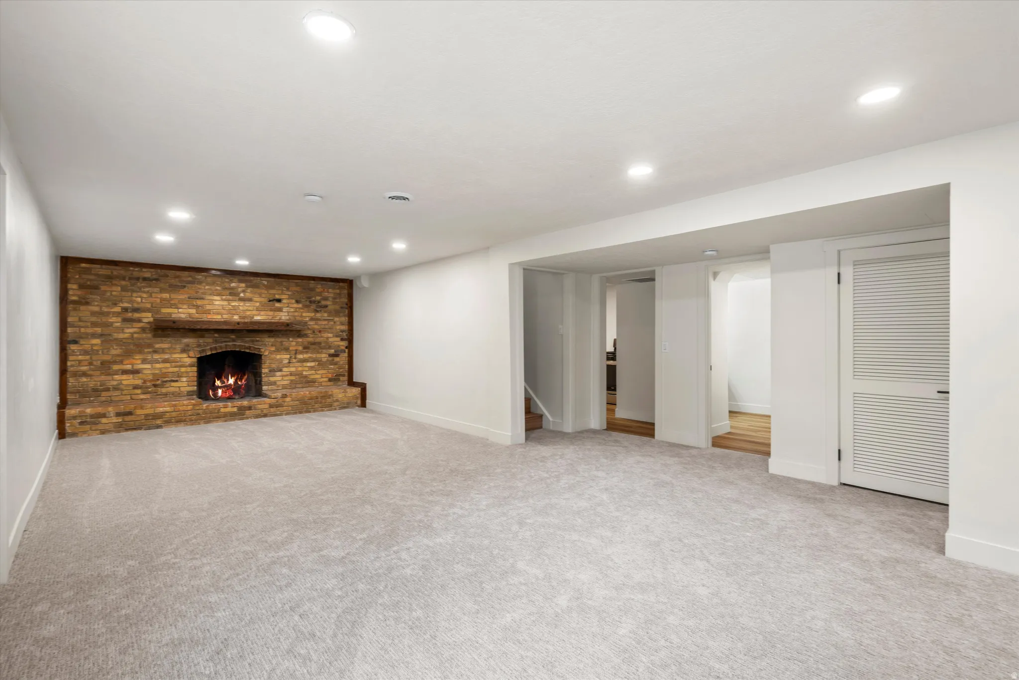 Spacious room featuring a brick fireplace with a wood mantle, recessed lighting, light-toned carpet, white walls, and louvered bi-fold doors