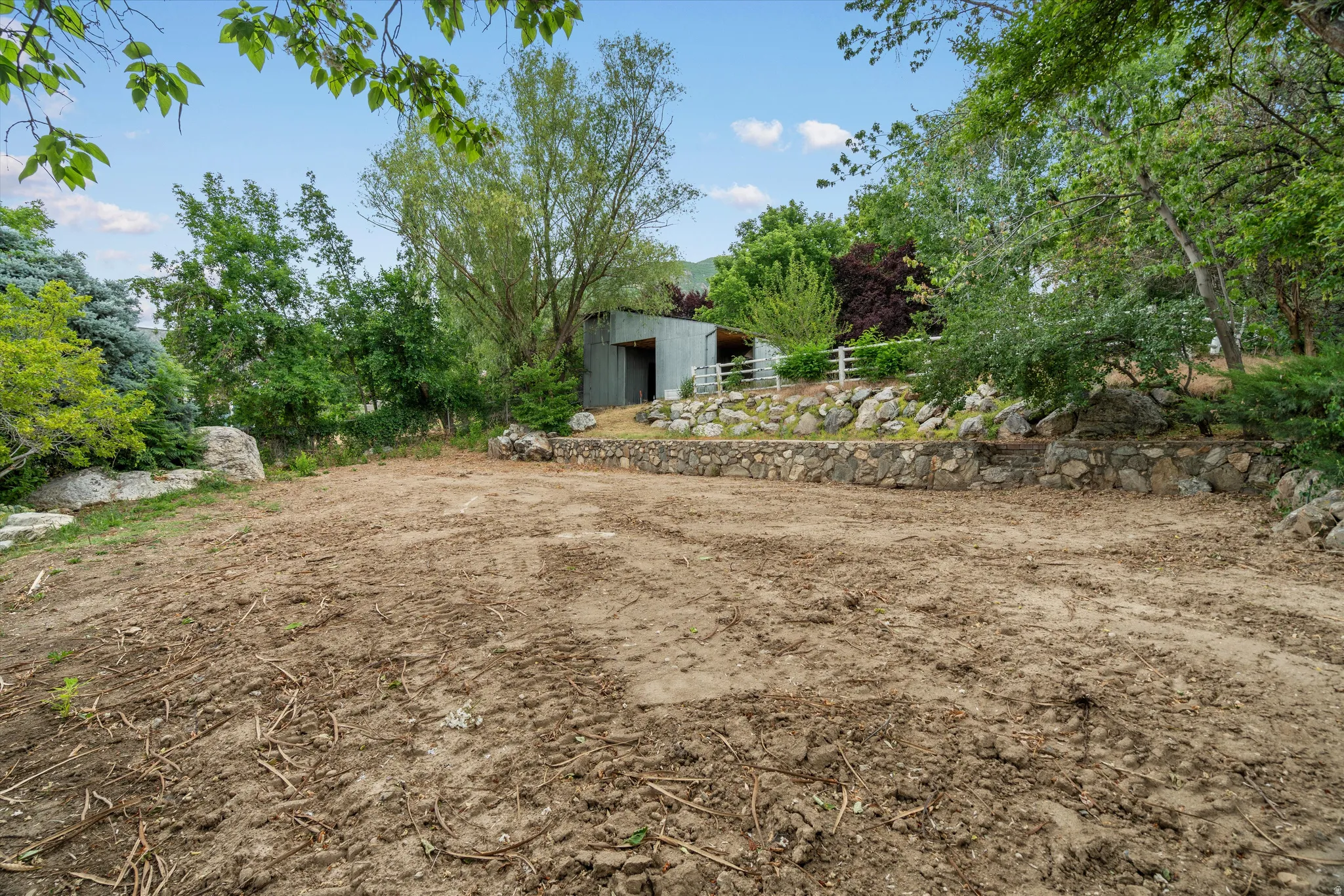 Cleared lot featuring a stone retaining wall, mature trees, and a utility shed