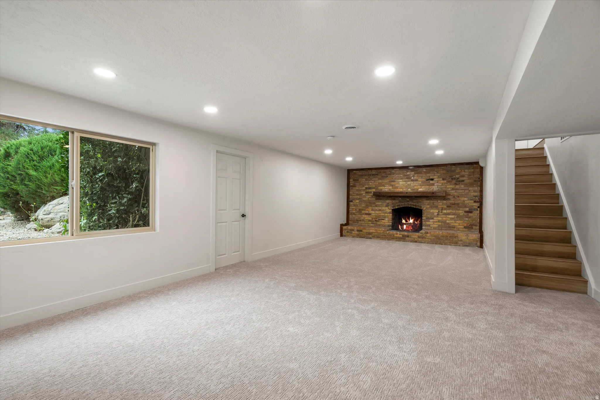 Expansive carpeted room featuring a stone-faced fireplace with a wood mantel, recessed lighting, and a window with exterior landscaping visible