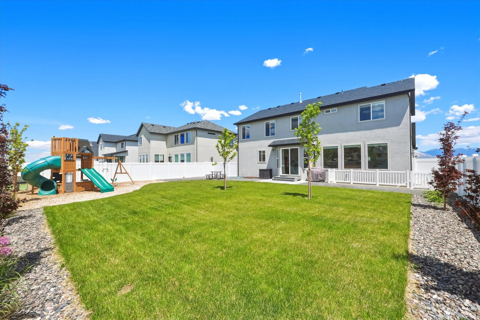 Expansive grassy yard with a white privacy fence, landscaping rocks, mature trees, and a ground-level patio with additional white fencing