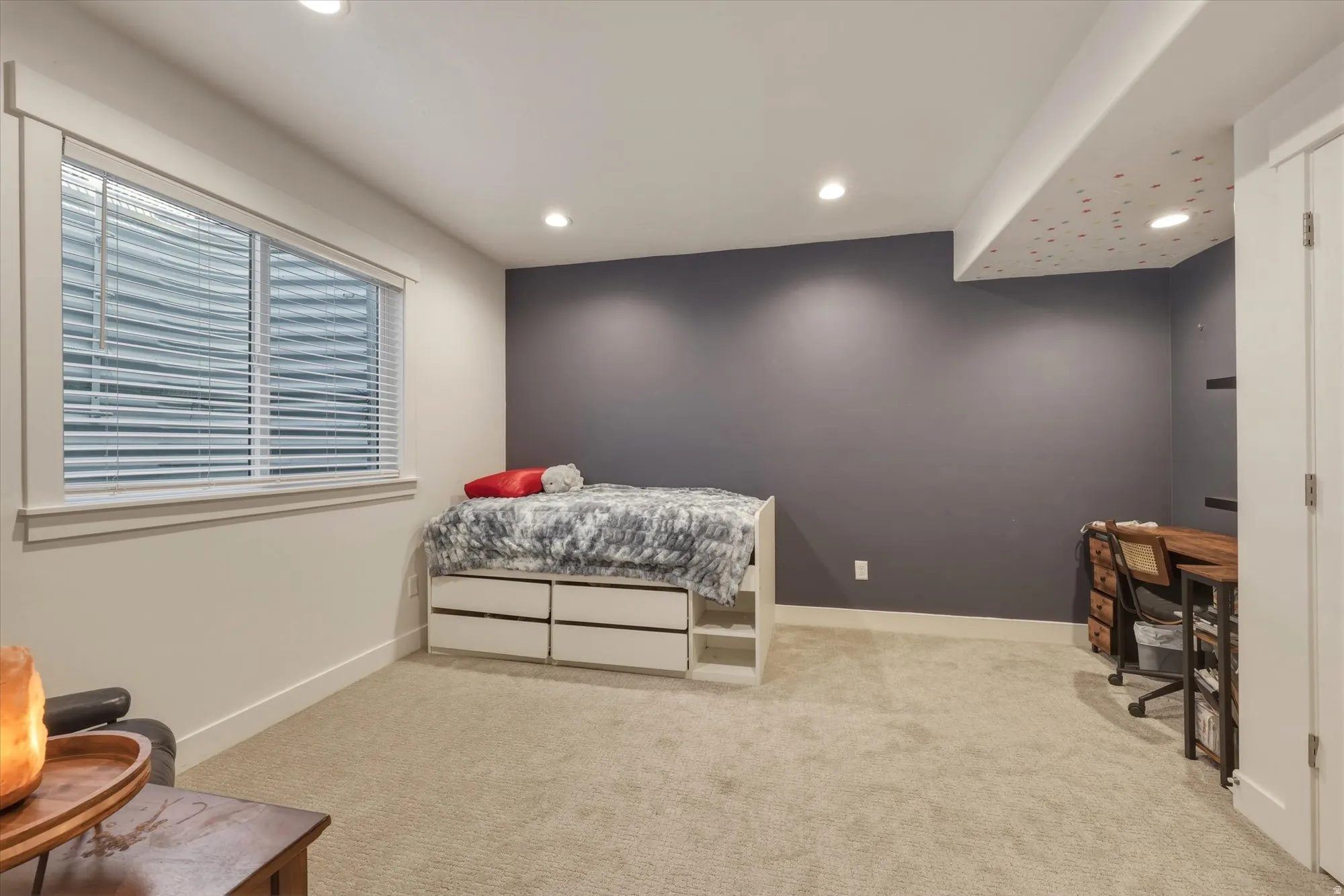 Spacious room featuring neutral carpeting, recessed ceiling lighting, a prominent window with white blinds, a contrasting accent wall, and white trim throughout
