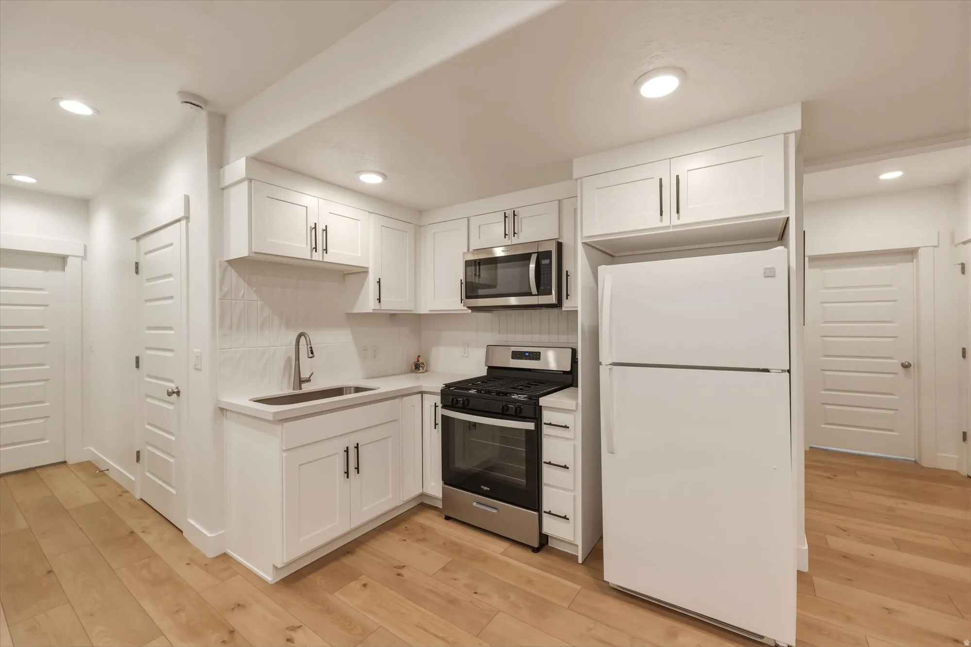 Kitchenette featuring white shaker-style cabinetry, a stainless steel gas range, a stainless steel microwave, a white refrigerator, a tile backsplash, and light wood-finish flooring