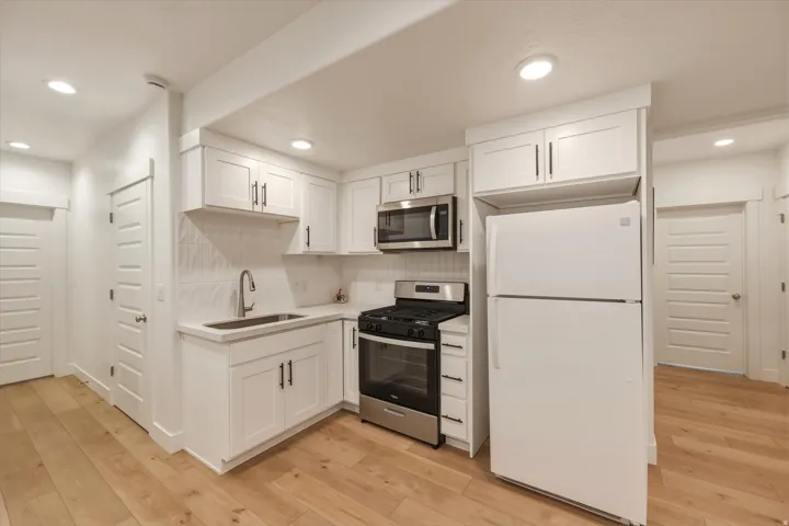 Kitchenette featuring white shaker-style cabinetry, a stainless steel gas range, a stainless steel microwave, a white refrigerator, a tile backsplash, and light wood-finish flooring