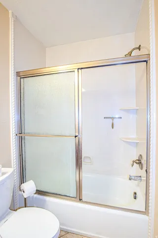 Second full bathroom featuring a shower-over-tub combination with sliding glass doors, a white acrylic tub surround with integrated shelving, and brass-tone fixtures