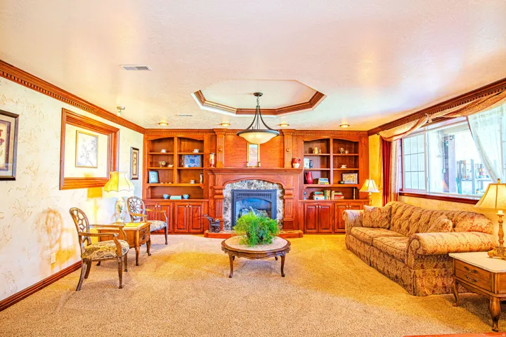 Living area featuring a stone fireplace with a wood mantle, built-in cabinetry and shelving, a coffered ceiling with a central chandelier, and large windows with transom details