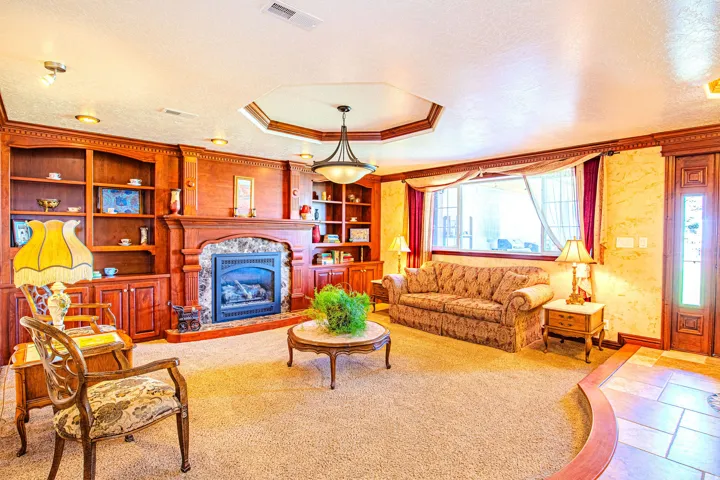 Stone-faced fireplace with a wood mantel and integrated built-in shelving, tray ceiling with a central chandelier, wood-finish flooring, and a large window