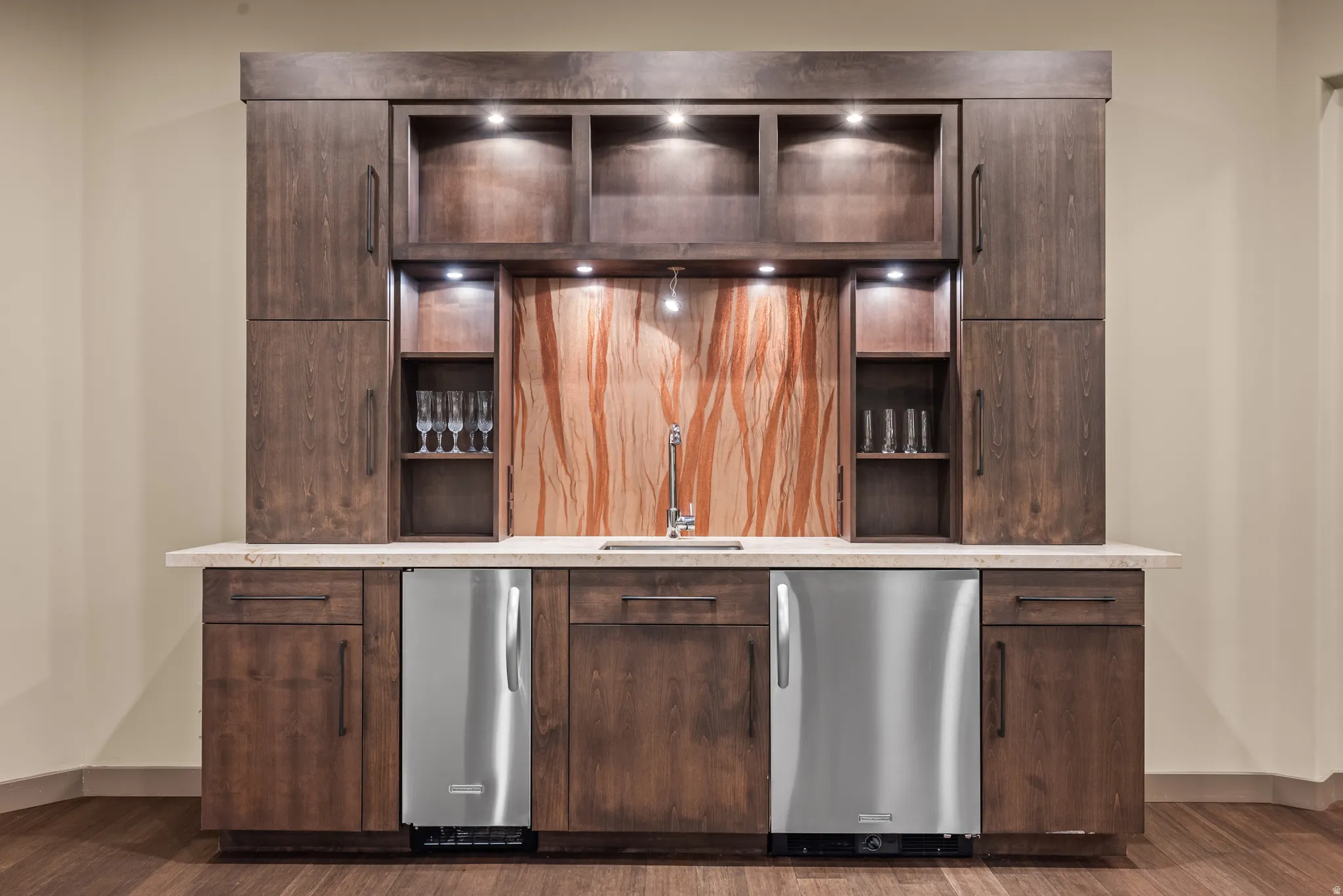 Built-in wet bar featuring dark wood-finish cabinetry, a patterned stone backsplash, undercabinet lighting, two stainless-steel beverage refrigerators, and a light-toned countertop