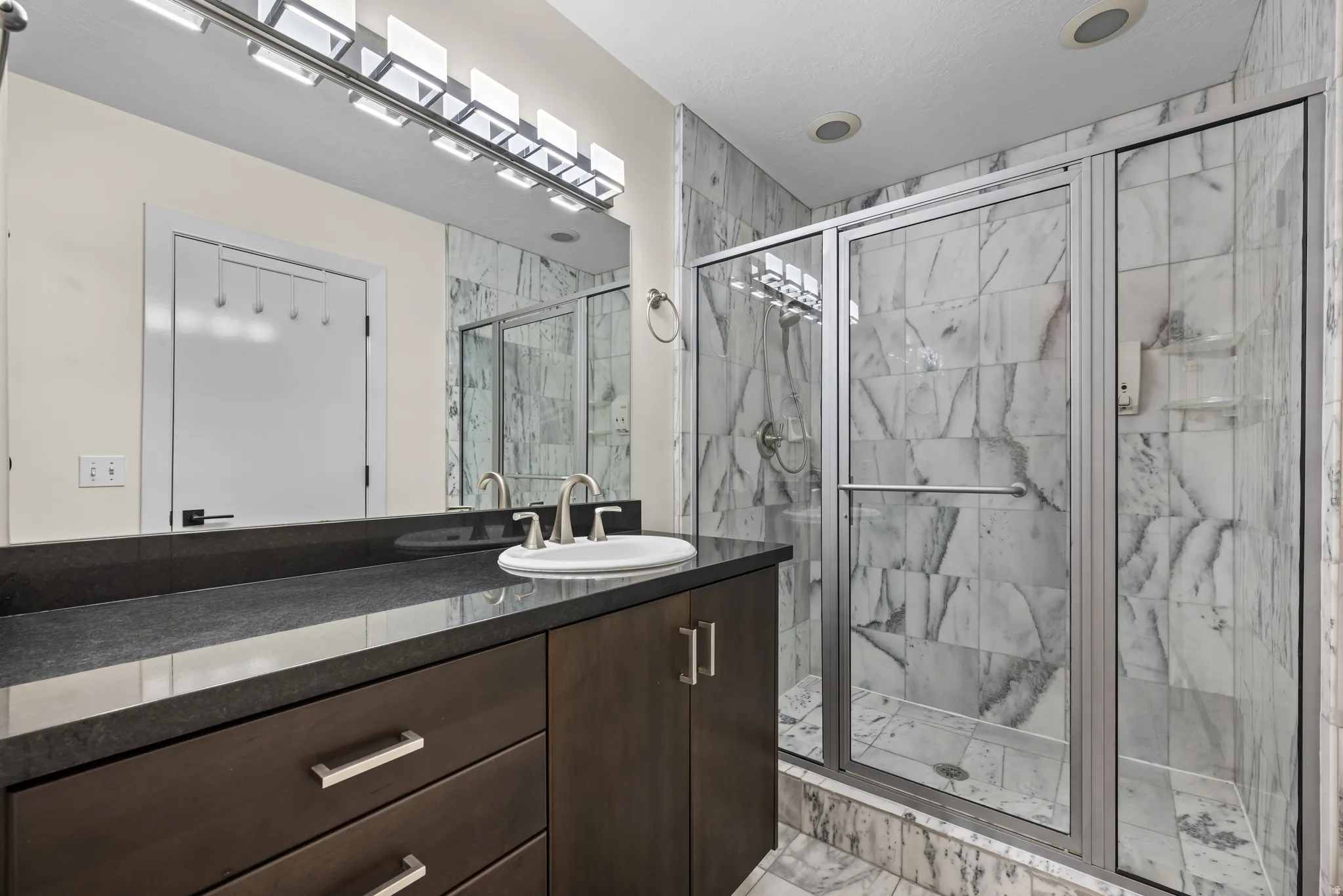 Bathroom featuring a spacious walk-in shower with glass enclosure, elegant marble-look wall tiling, a dark wood-finish vanity with a single basin, a large mirror, and contemporary lighting fixtures