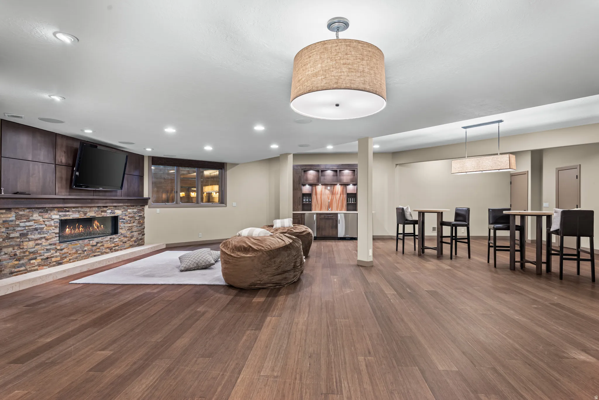 Spacious room featuring wood-finish flooring, a stone-clad fireplace with built-in cabinetry, recessed lighting, and a wet bar with integrated appliances