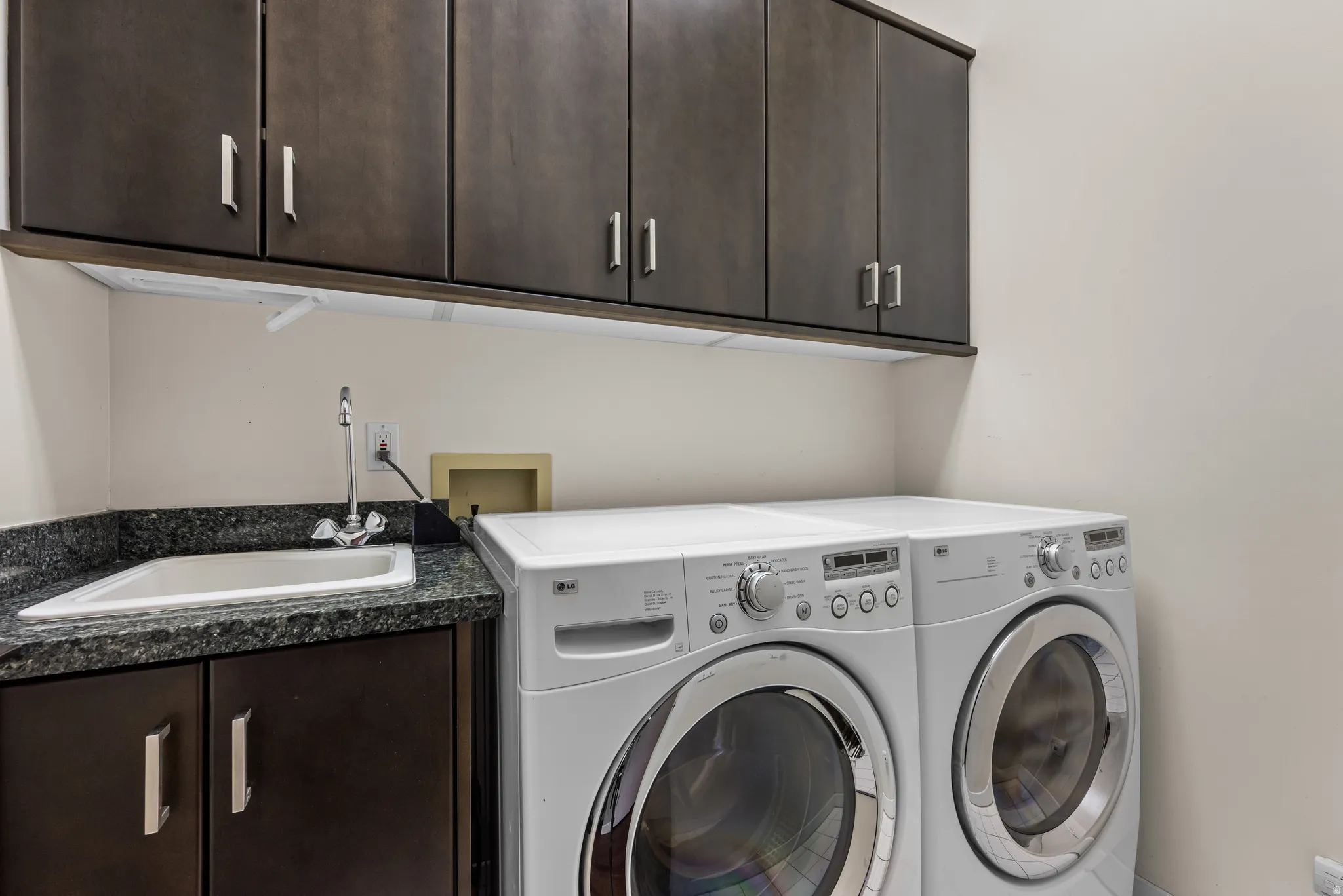 Utility room featuring dark wood-finish cabinetry, a laundry sink with a chrome faucet, and a stone-look countertop