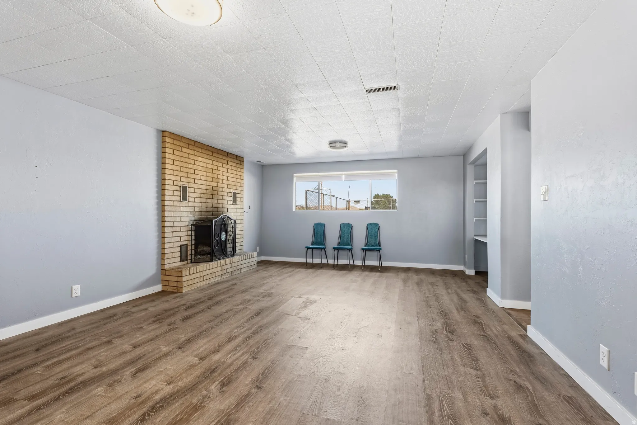 Open living area featuring wood-finish flooring, a brick-faced fireplace, a large window, light blue walls, and built-in shelving