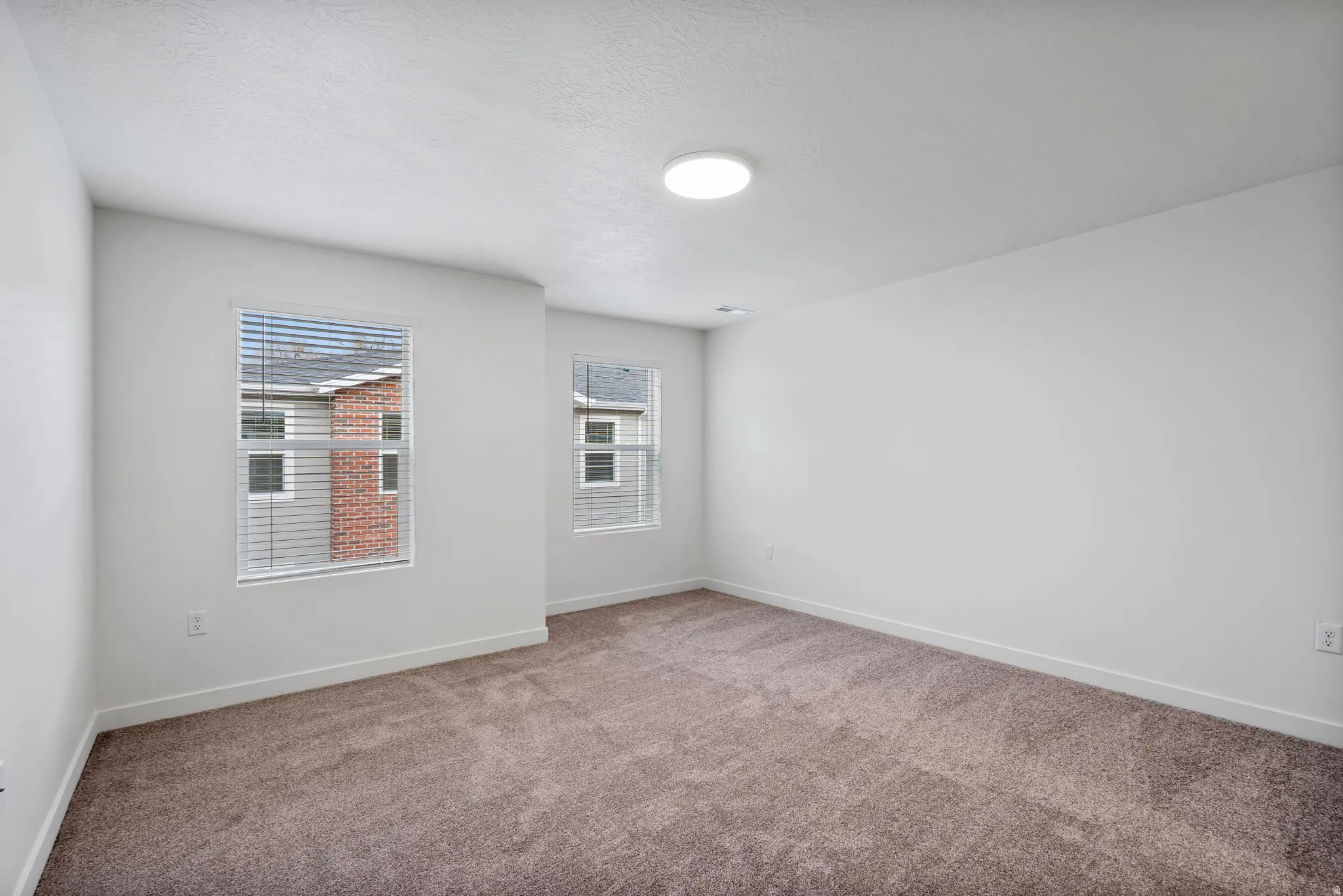 Neutral tone carpeting in a room featuring two windows with horizontal blinds and a ceiling-mounted light fixture