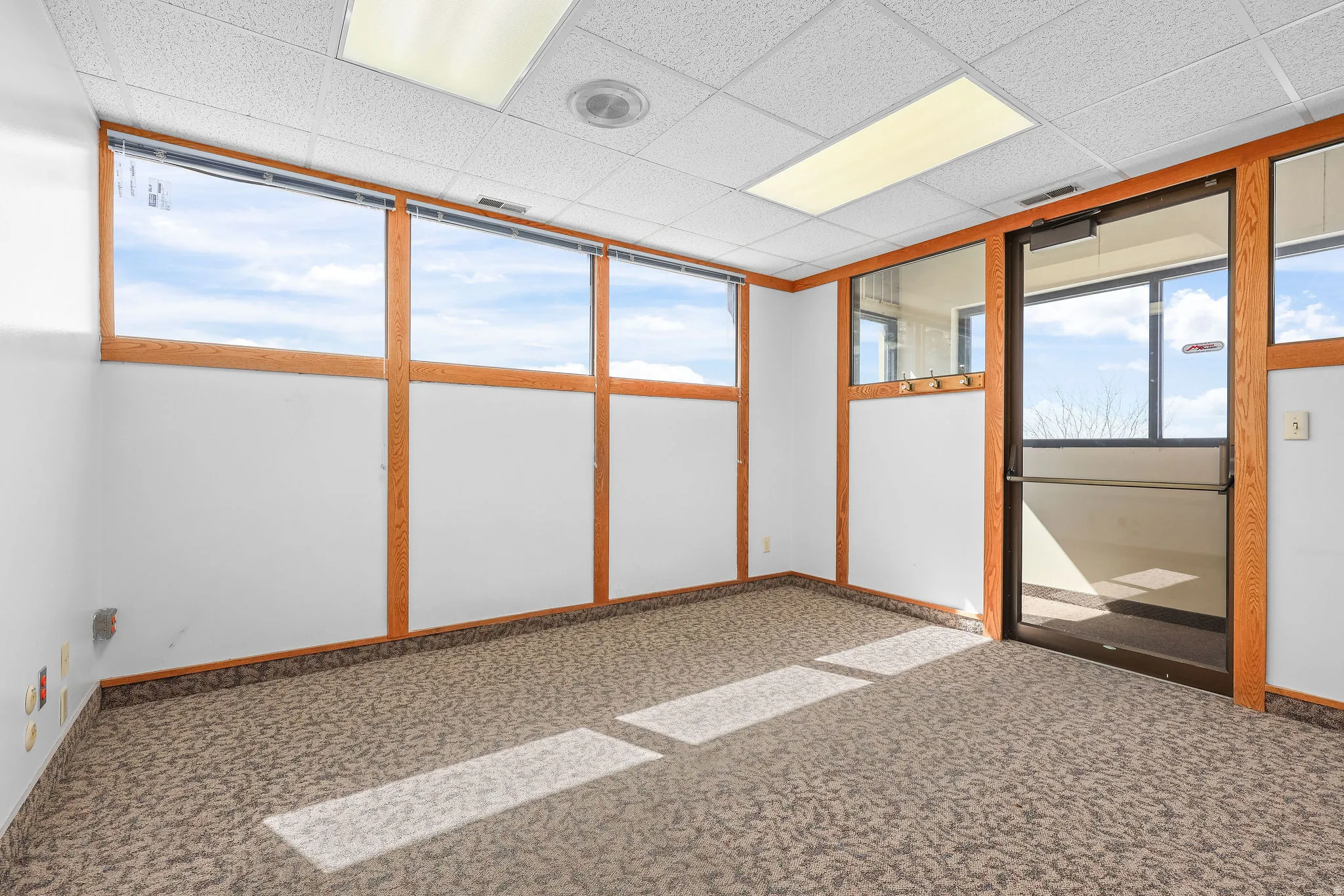 Commercial space featuring expansive window-lined walls, a drop ceiling with integrated lighting, patterned carpet flooring, and a glass-paneled entry door with a push bar