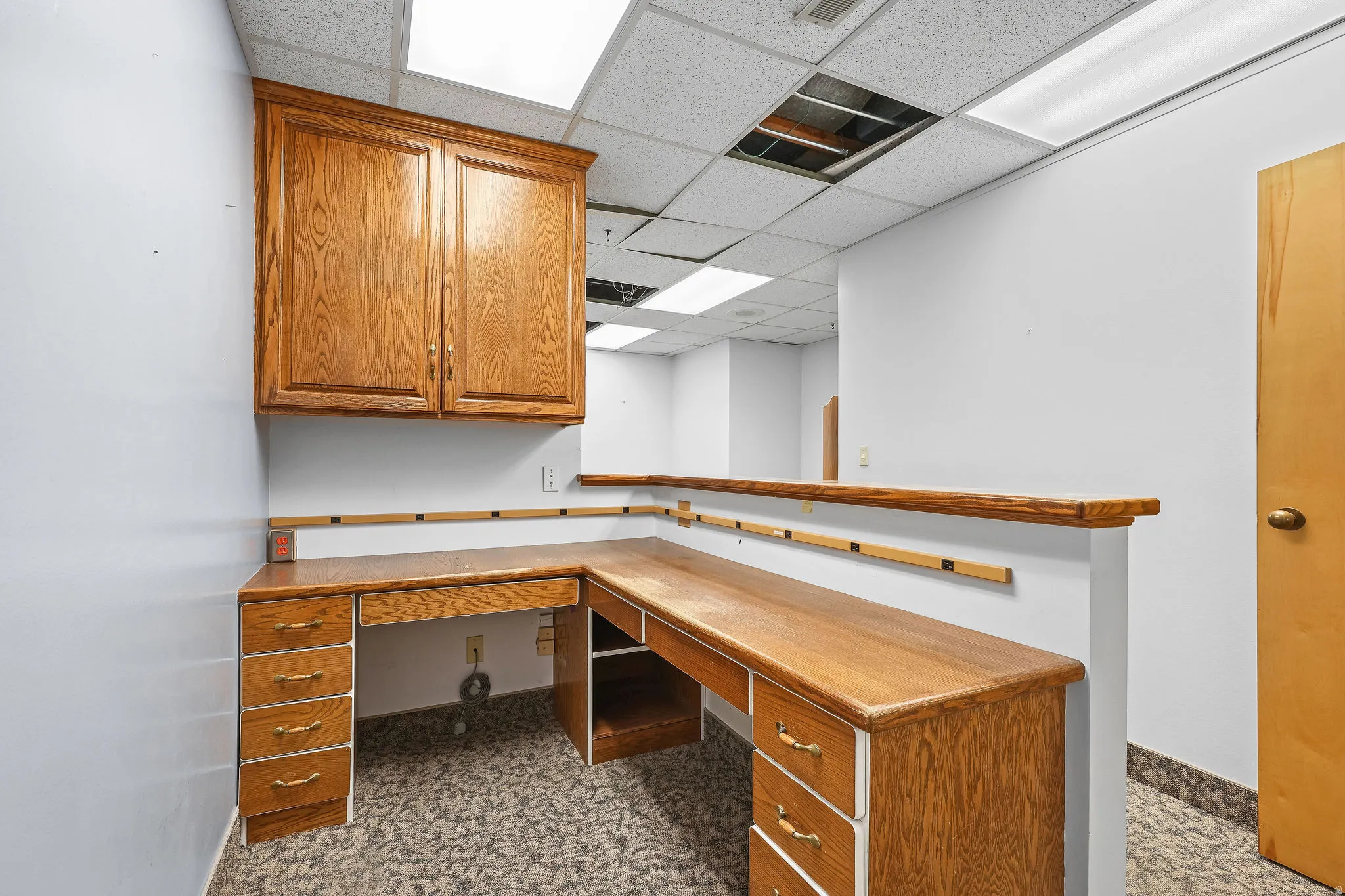 Built-in oak desk with drawer storage and upper cabinetry, accented by white walls and a drop ceiling