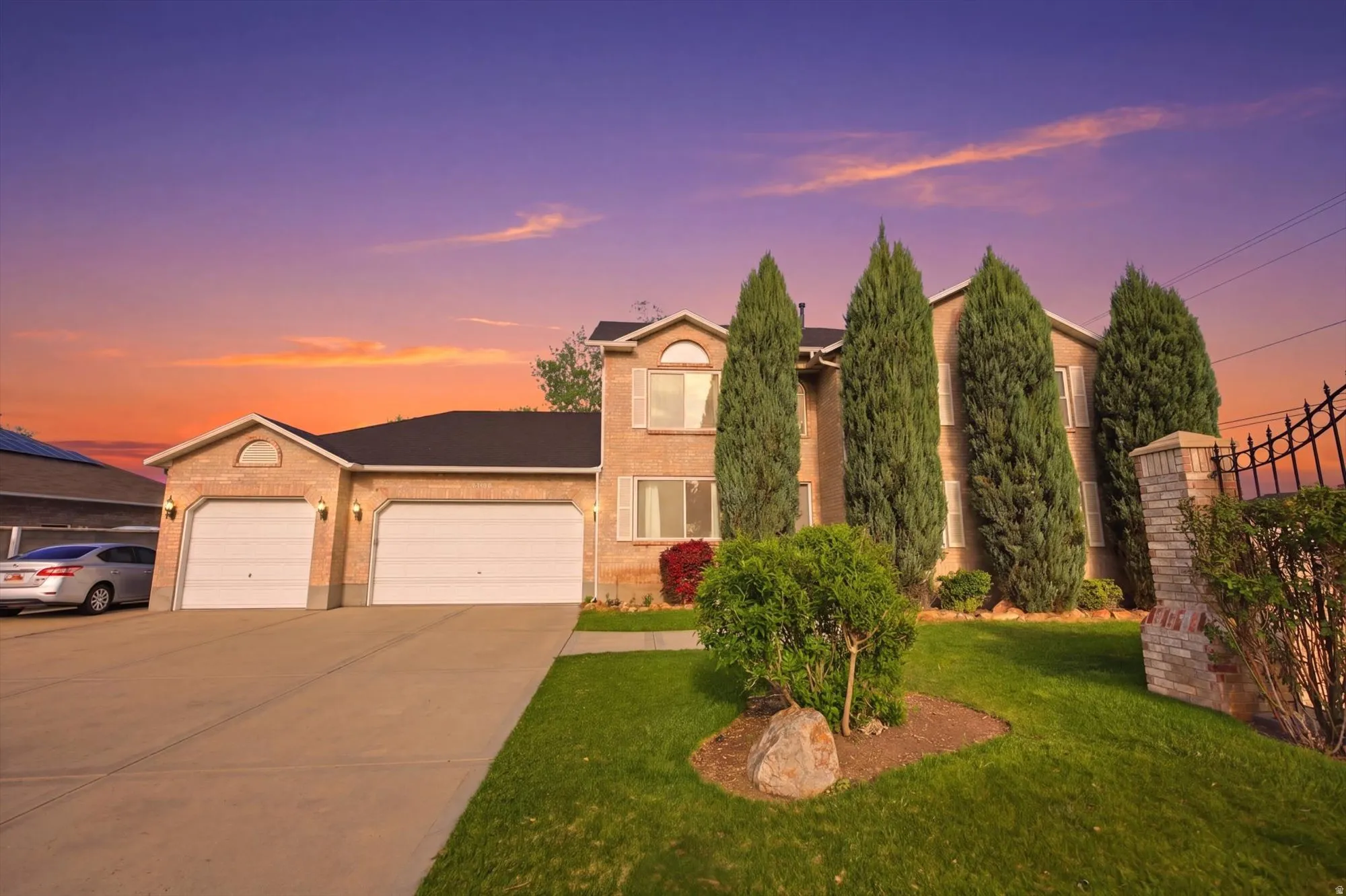 Brick-front residence featuring a two-car garage, mature landscaping, a paved driveway, and a decorative wrought iron fence